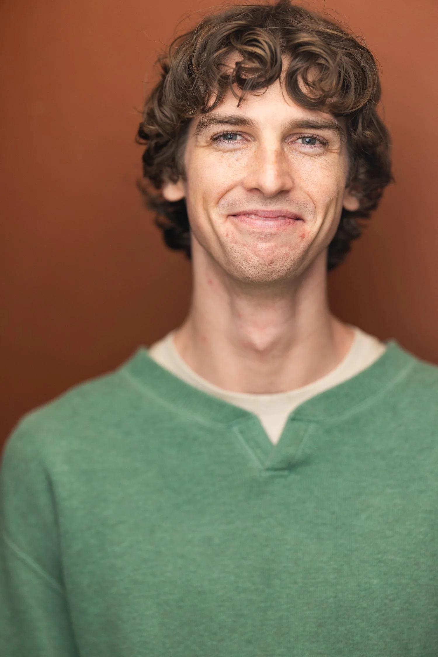 A young man with curly brown hair and blue eyes, wearing a green sweatshirt, smiling slightly in front of a brown background.