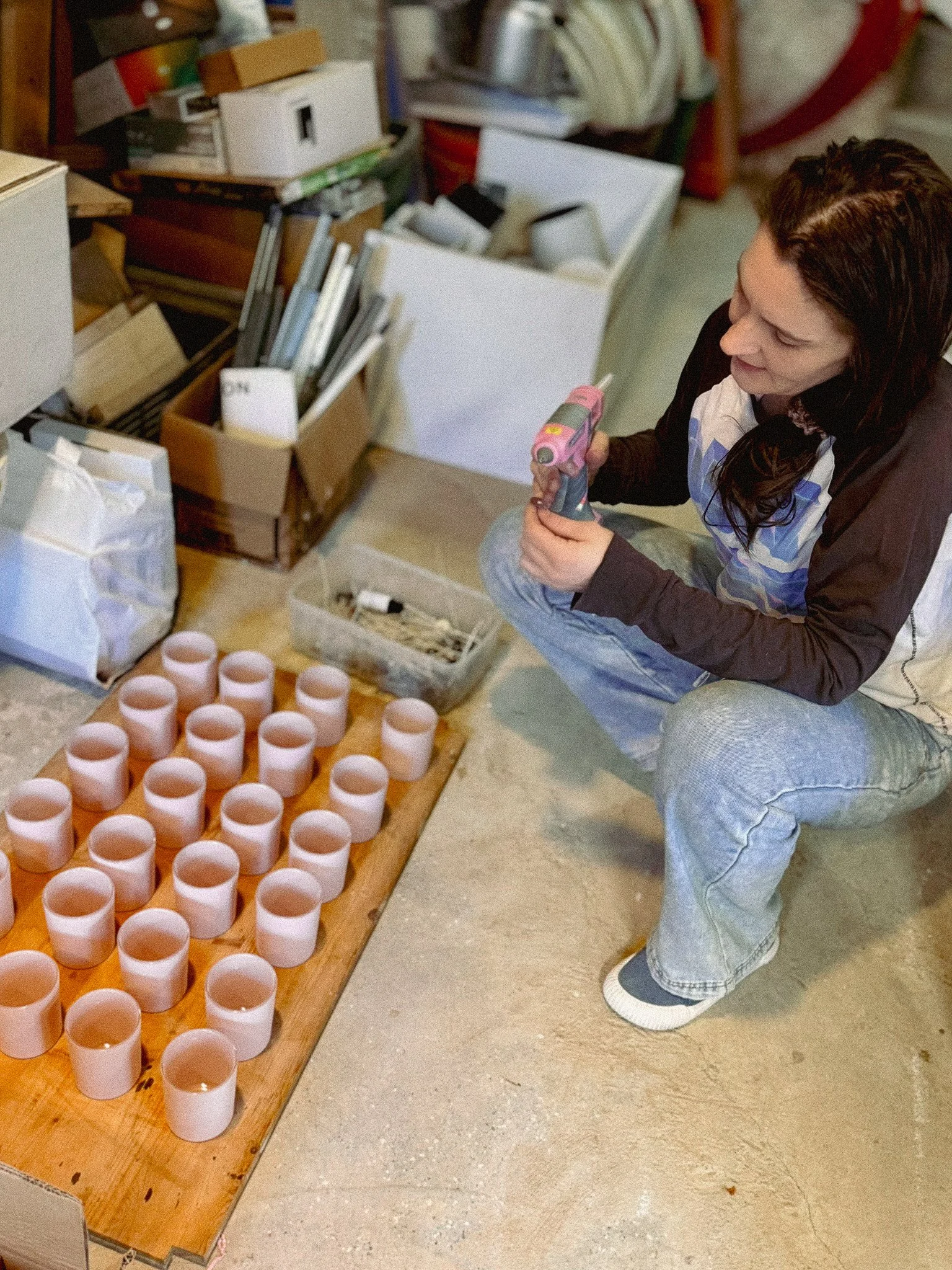 A person in casual clothing is sitting on the floor of a storage room, holding a pink cordless drill. There are several pink cups arranged on a wooden table in front of them, and boxes filled with various items, including white cups and rolled-up paper towels, are visible around them.