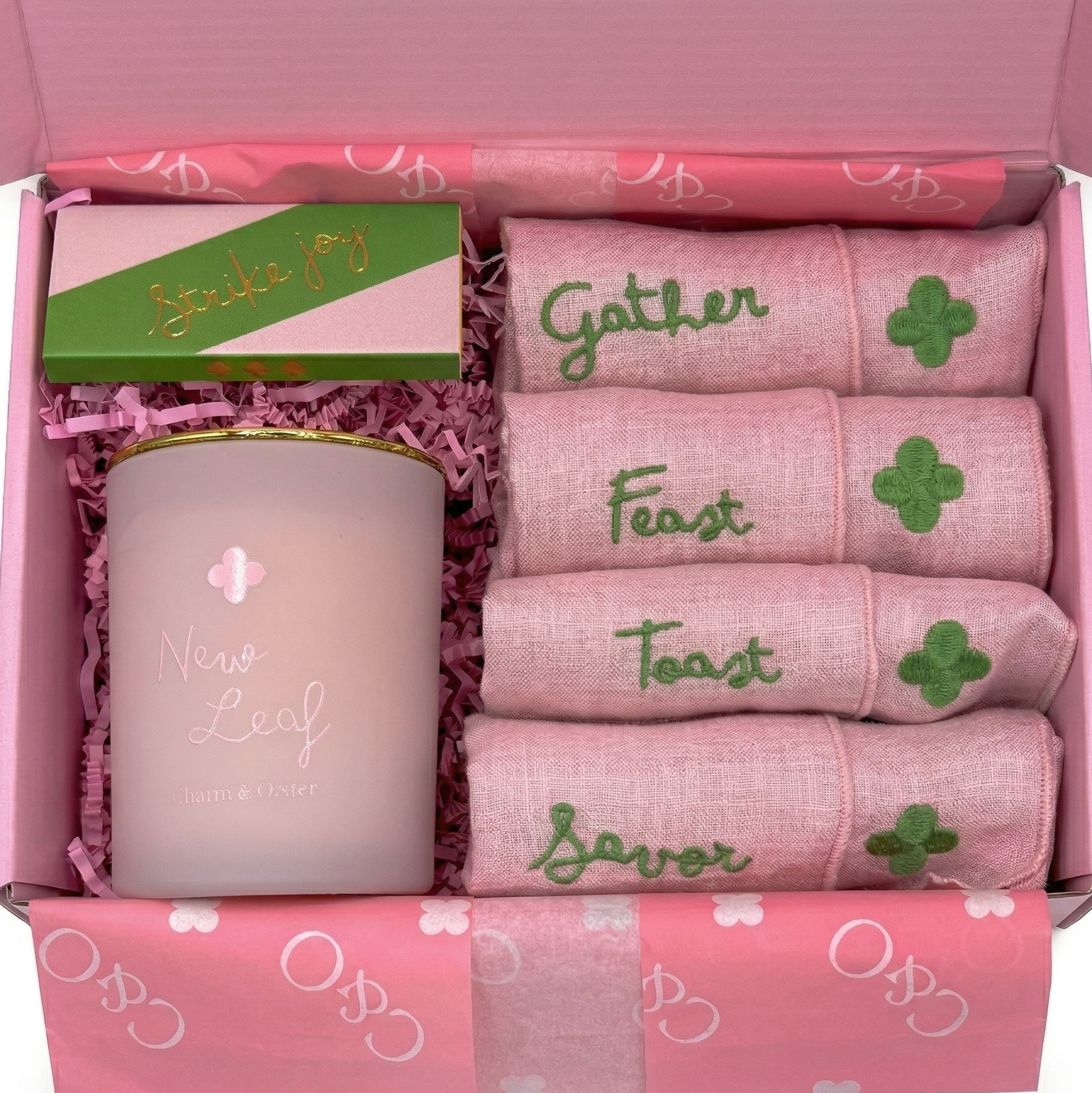 Pink gift box with accessories inside, including a pink candle with a clover emblem and matching embroidered pink towels with green words 'Gather', 'Feast', and 'Celebrate' and green clover symbols.