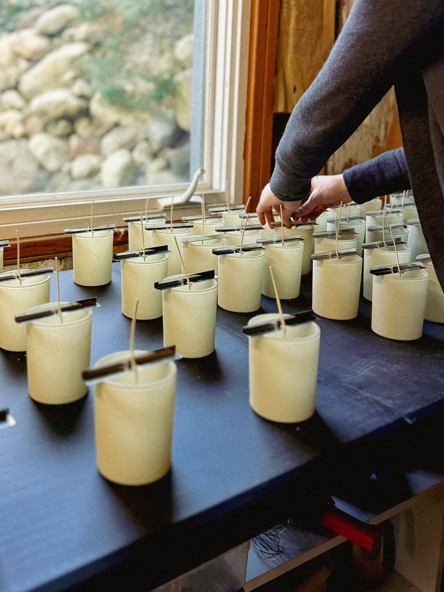Several white candles with black labels and wicks arranged on a black table near a window made of wood, with rocks and greenery visible outside.