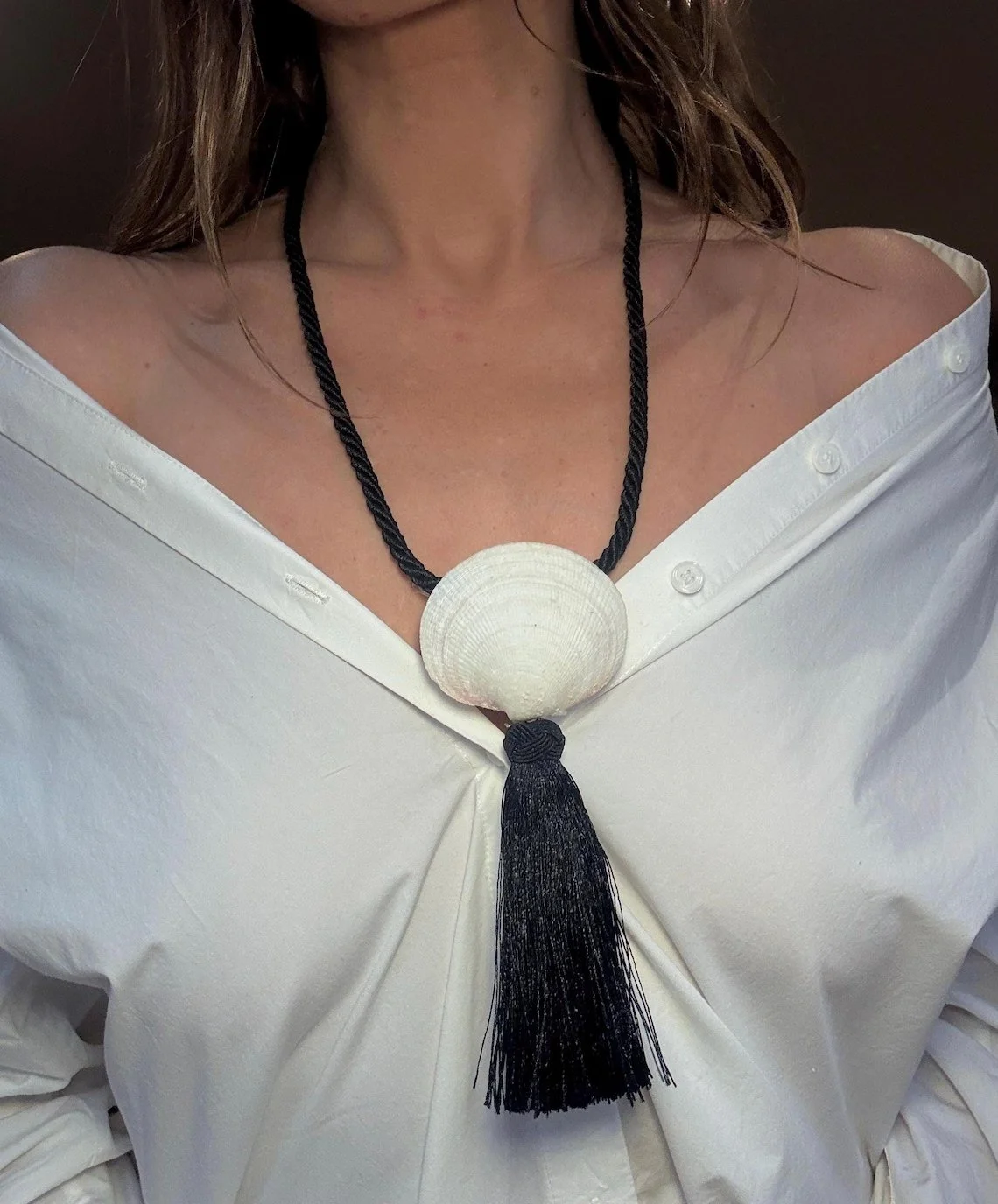 Close-up of a woman wearing a white off-shoulder blouse and a black and white statement necklace with a large shell and black tassel.