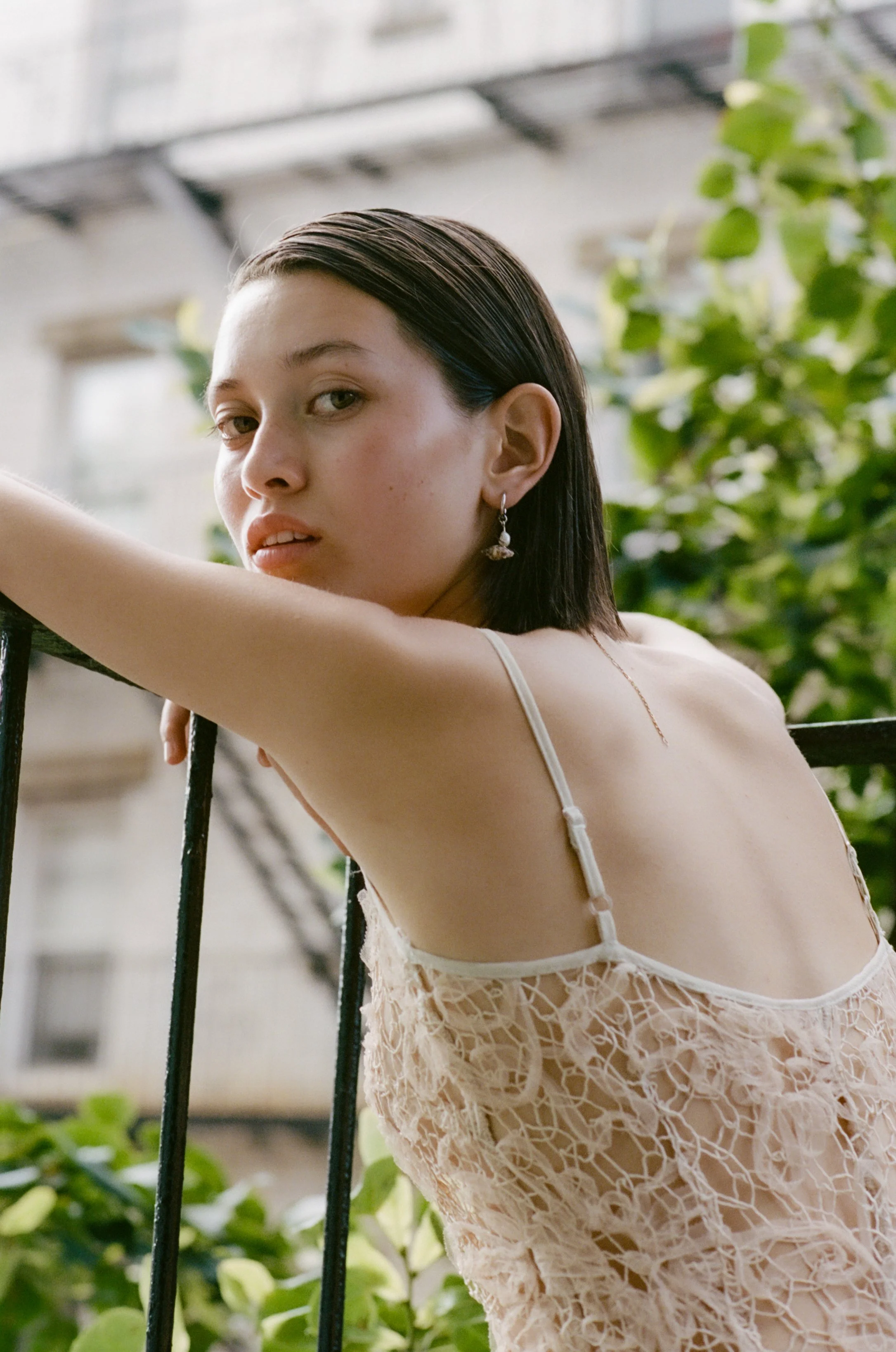 A young woman with dark hair, wearing a beige lace dress and earrings, leaning on a railing on a balcony with greenery in the background.