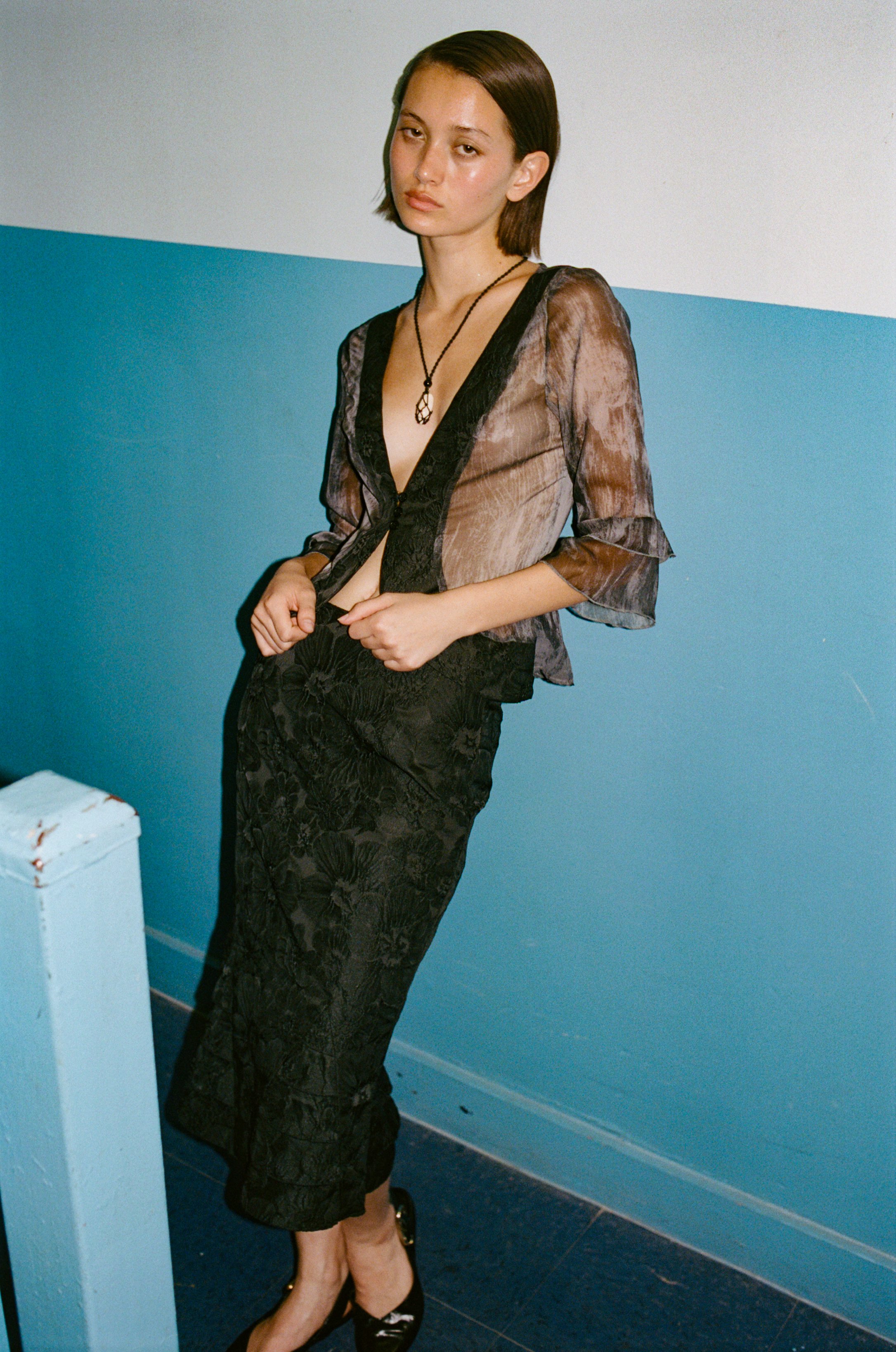 Young woman with short brown hair in sheer, patterned blouse and black patterned skirt, standing against a two-tone wall with a blue lower half and white upper half.