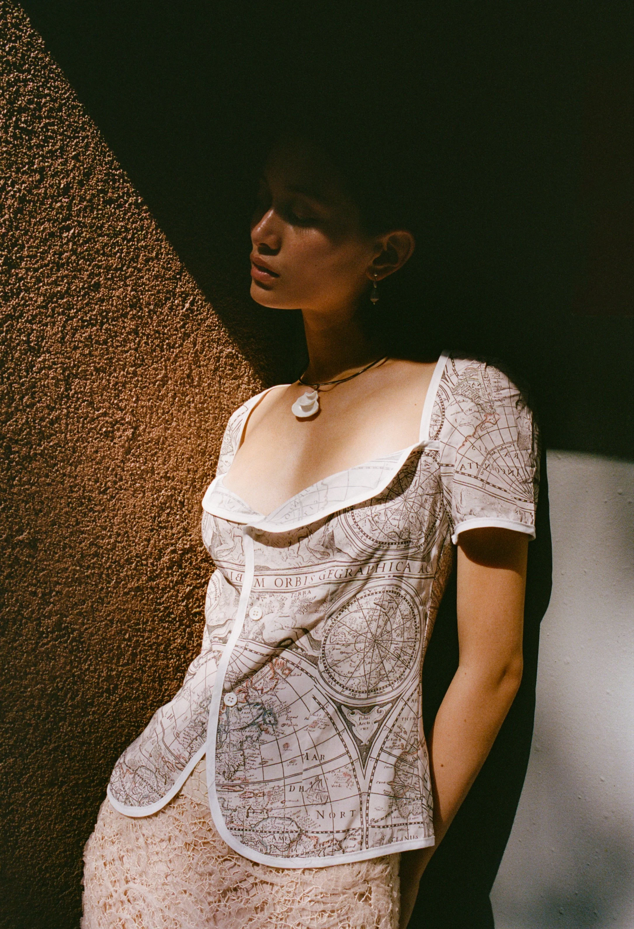A woman standing indoors against a wall, wearing a vintage map-printed shirt with a collar and lace skirt. She has short dark hair, earrings, and a choker necklace with a shell pendant. She looks down with a calm expression, partially shadowed by dramatic lighting.