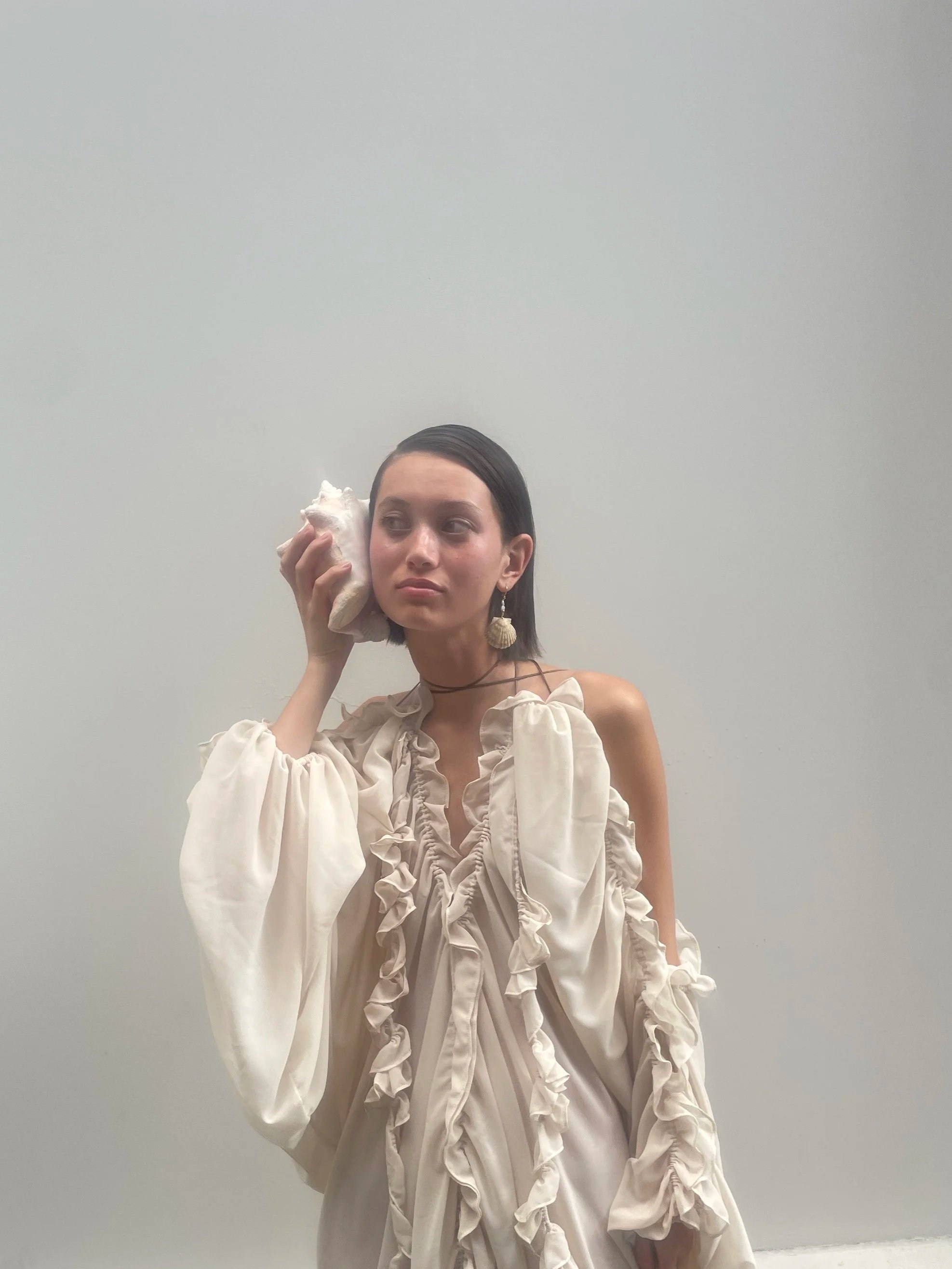 A young woman with dark hair and earrings stands against a plain white wall, holding a cloth to her ear as if listening, wearing a ruffled, cream-colored dress with off-the-shoulder sleeves.