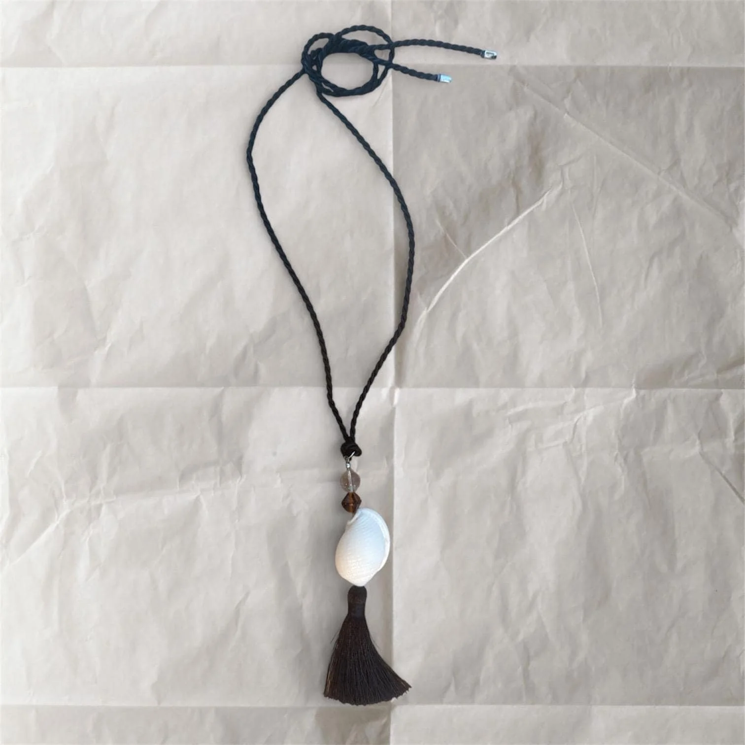 A necklace with a black cord, a white seashell pendant, small brown and clear beads, and a dark tassel at the end, laid on a crumpled white paper background.