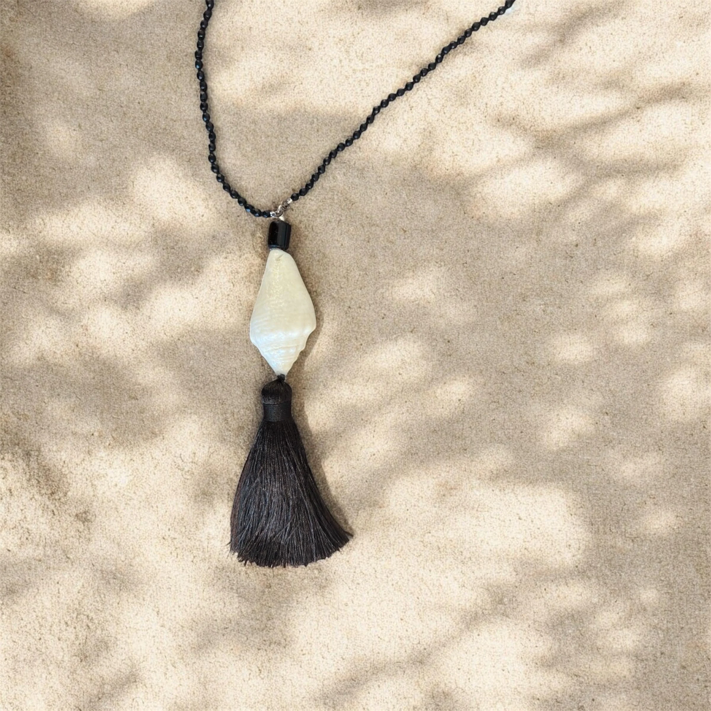 Necklace with a black beaded chain, large white shell pendant, and black tassel, resting on sandy surface with shadows.
