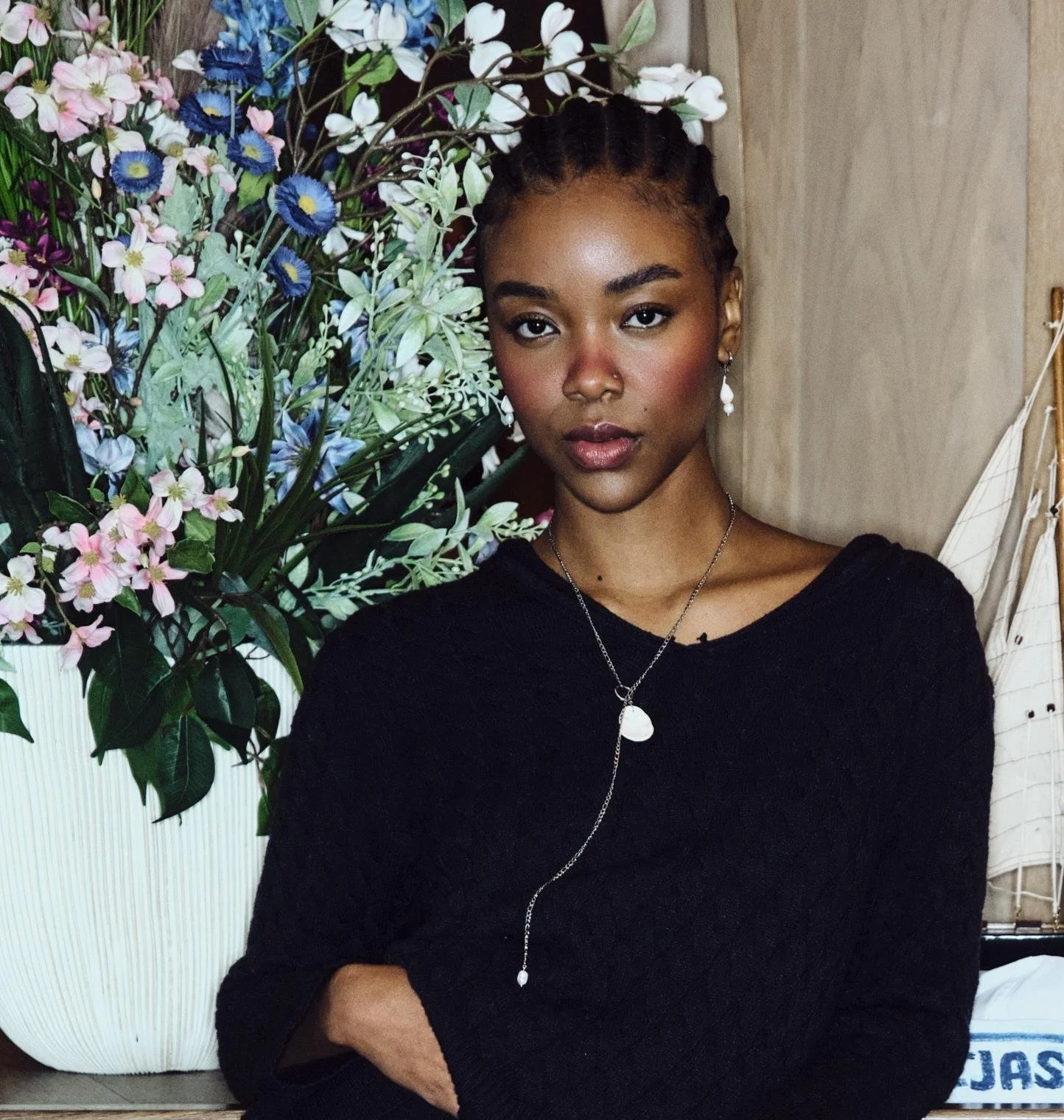 A young woman with braided hair and dark skin posing indoors in front of a large bouquet of multicolored flowers. She is wearing a black sweater, pearl earrings, and a long silver necklace with a pendant.