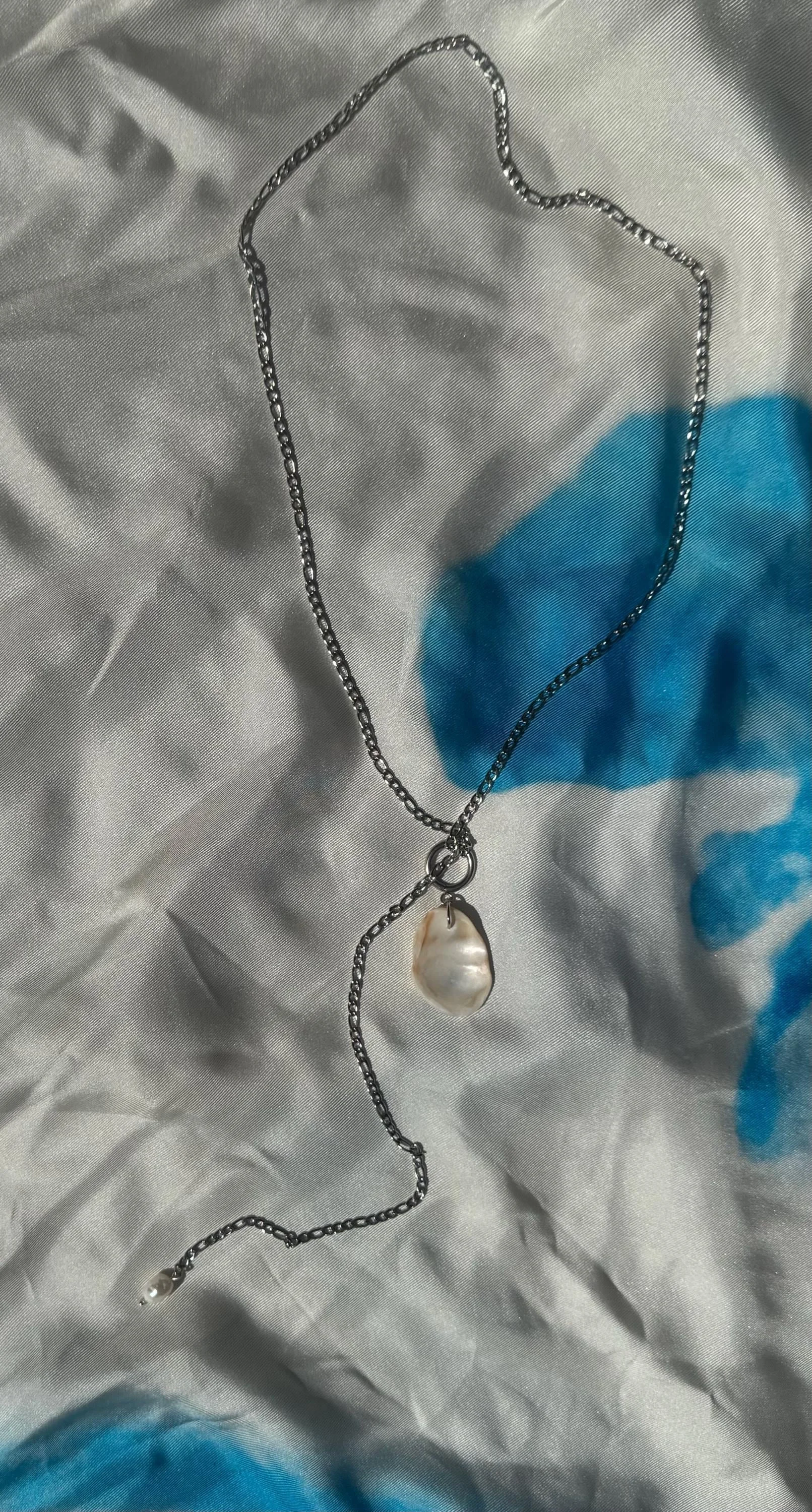 Silver chain necklace with a large, irregularly shaped pearl pendant resting on crinkled fabric with blue and white colors.