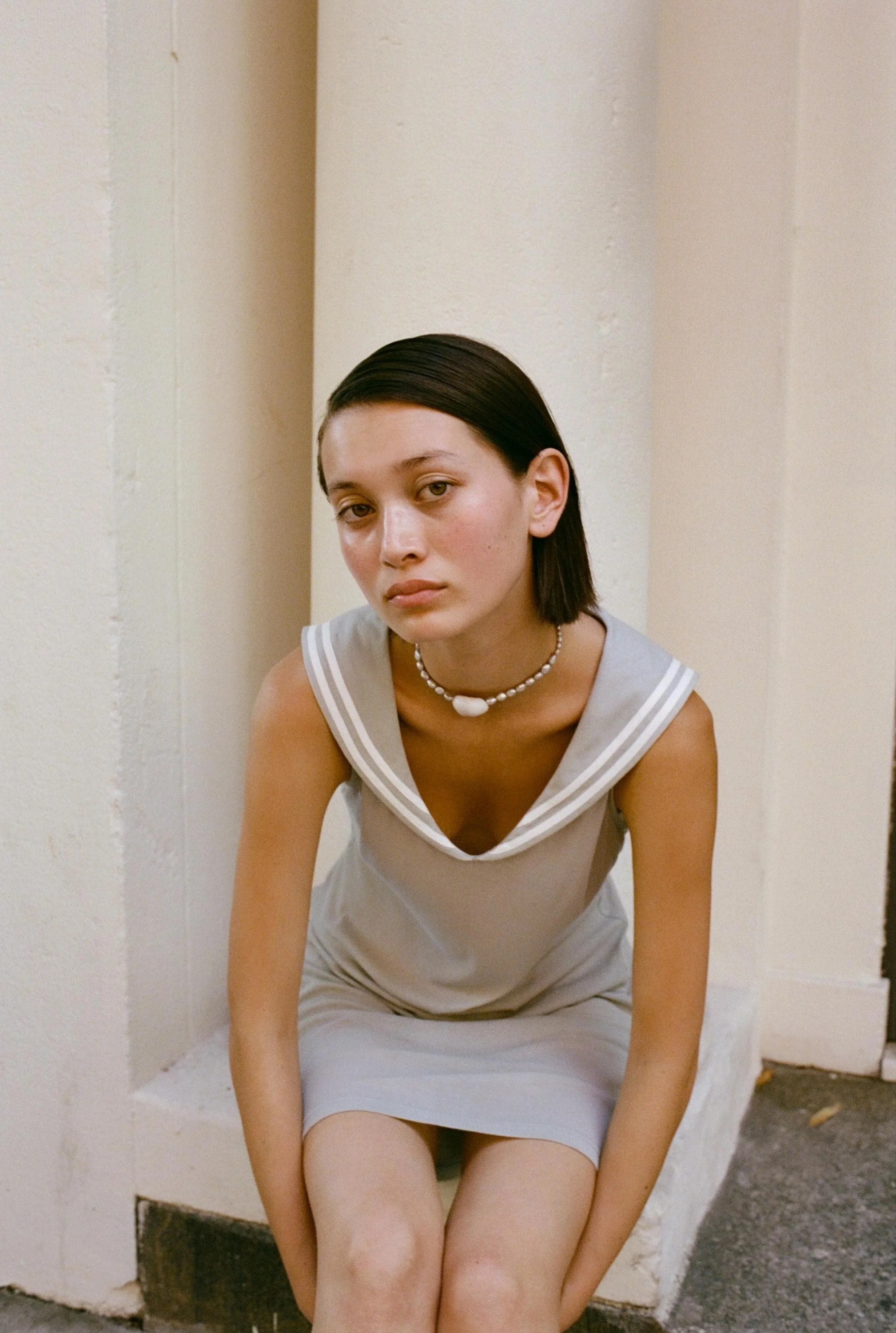 A young woman with dark hair sitting on a small ledge against a plain white wall, wearing a light-colored sleeveless top with a sailor collar and a pearl necklace.