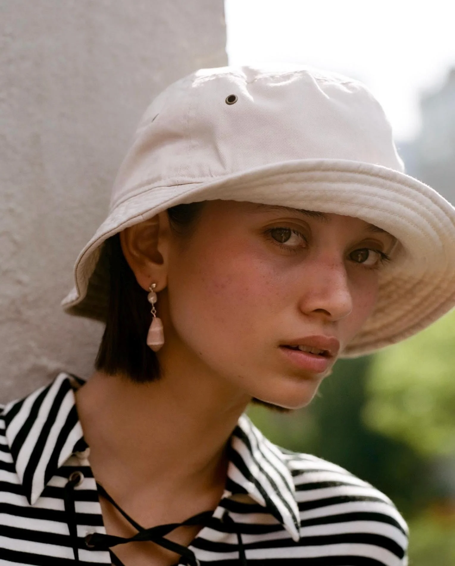 A young woman wearing a white bucket hat, black and white striped shirt, and dangling earring, looking at the camera with a neutral expression.