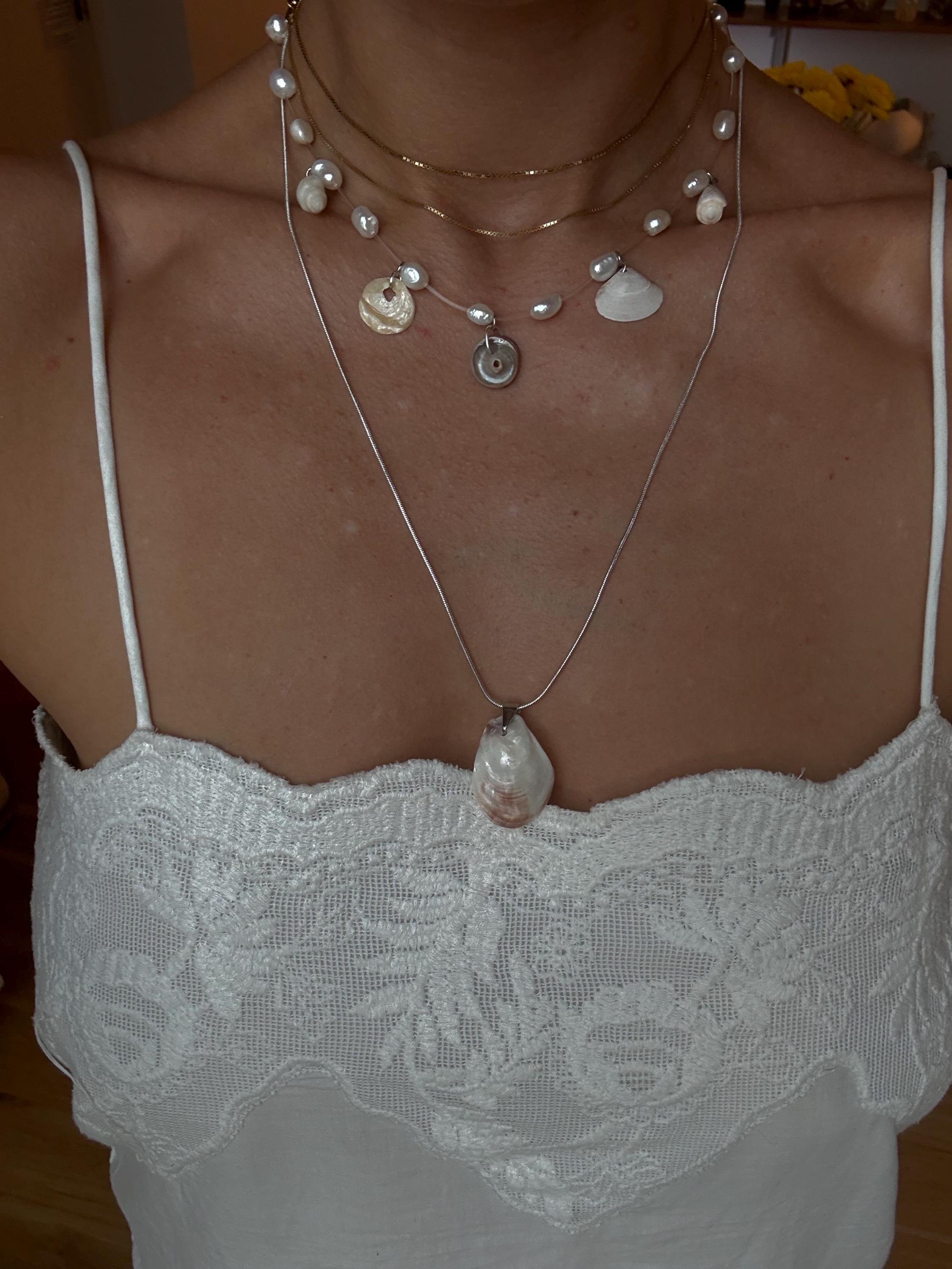 A woman wearing layered pearl and shell necklaces with a large shell pendant, and a white lace top with spaghetti straps.