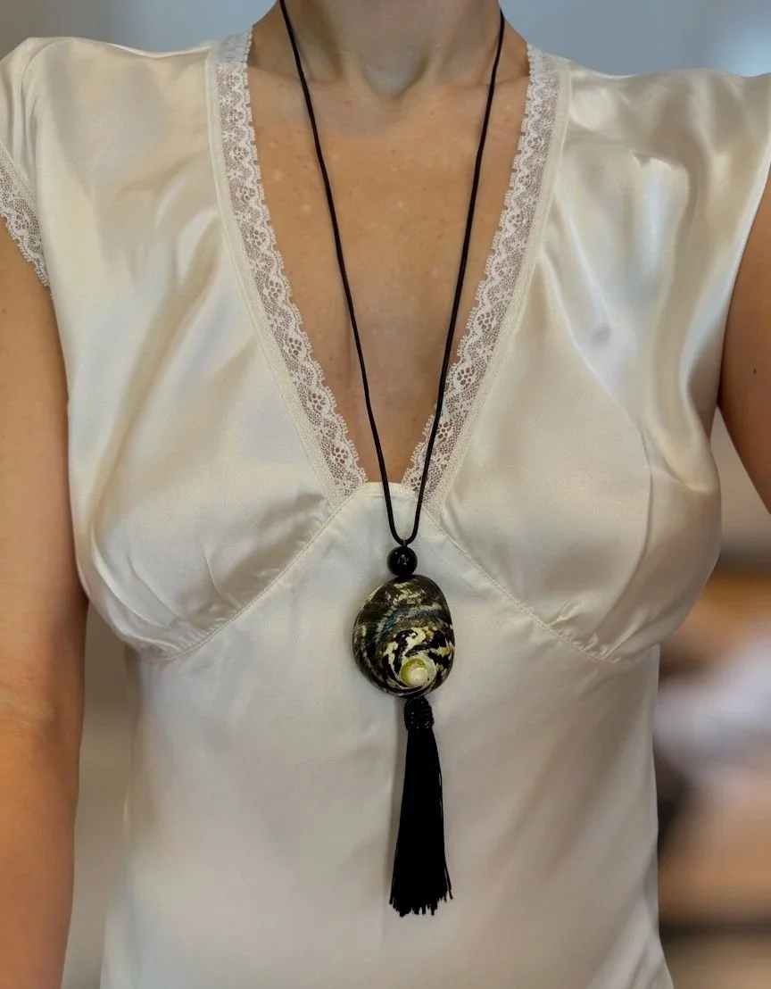 Woman wearing a satin white top with lace trim and a long black necklace with a large, patterned, oval pendant and black tassel.