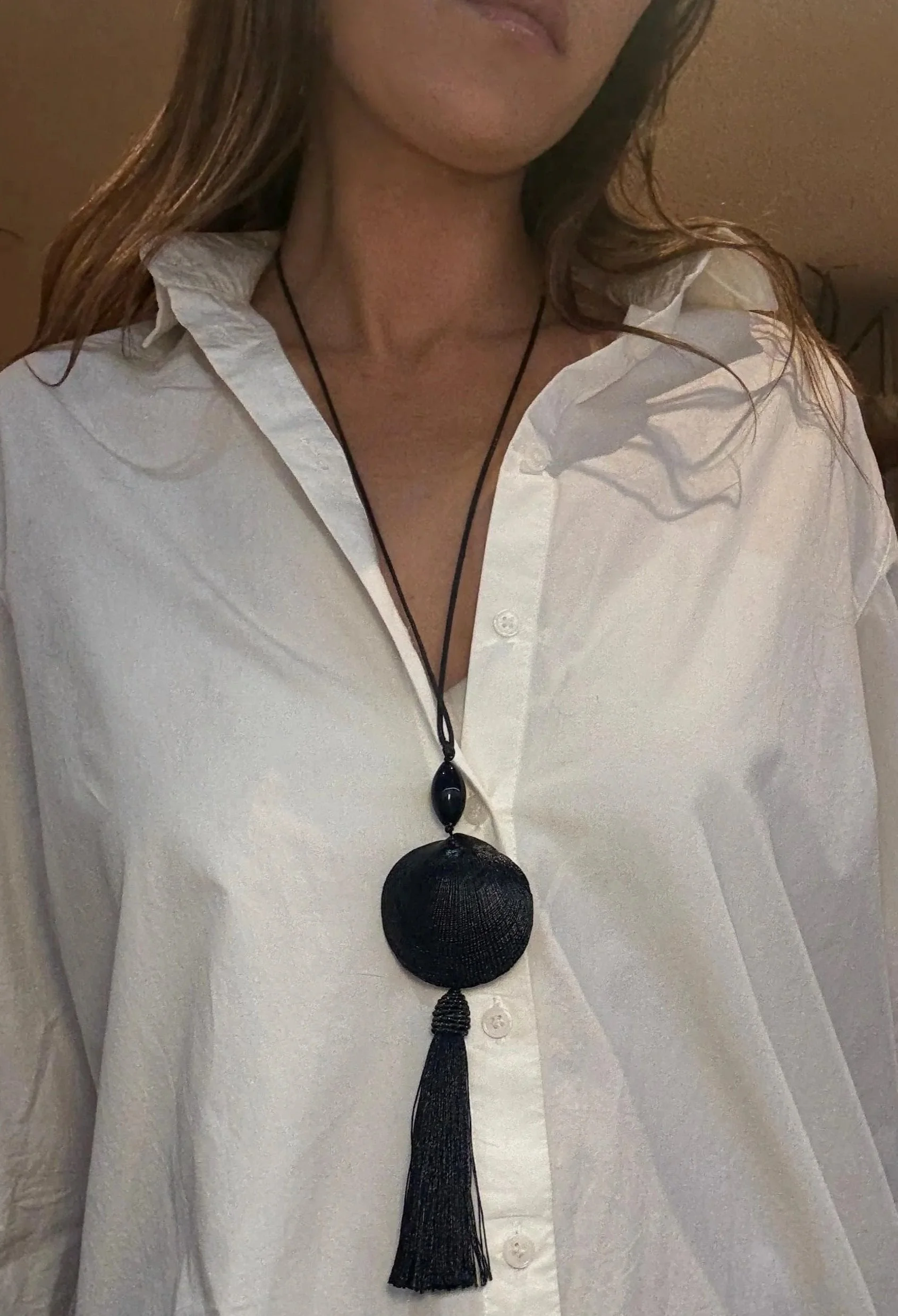 A woman wearing a white button-up shirt and a long black necklace with a large circular pendant and tassel.