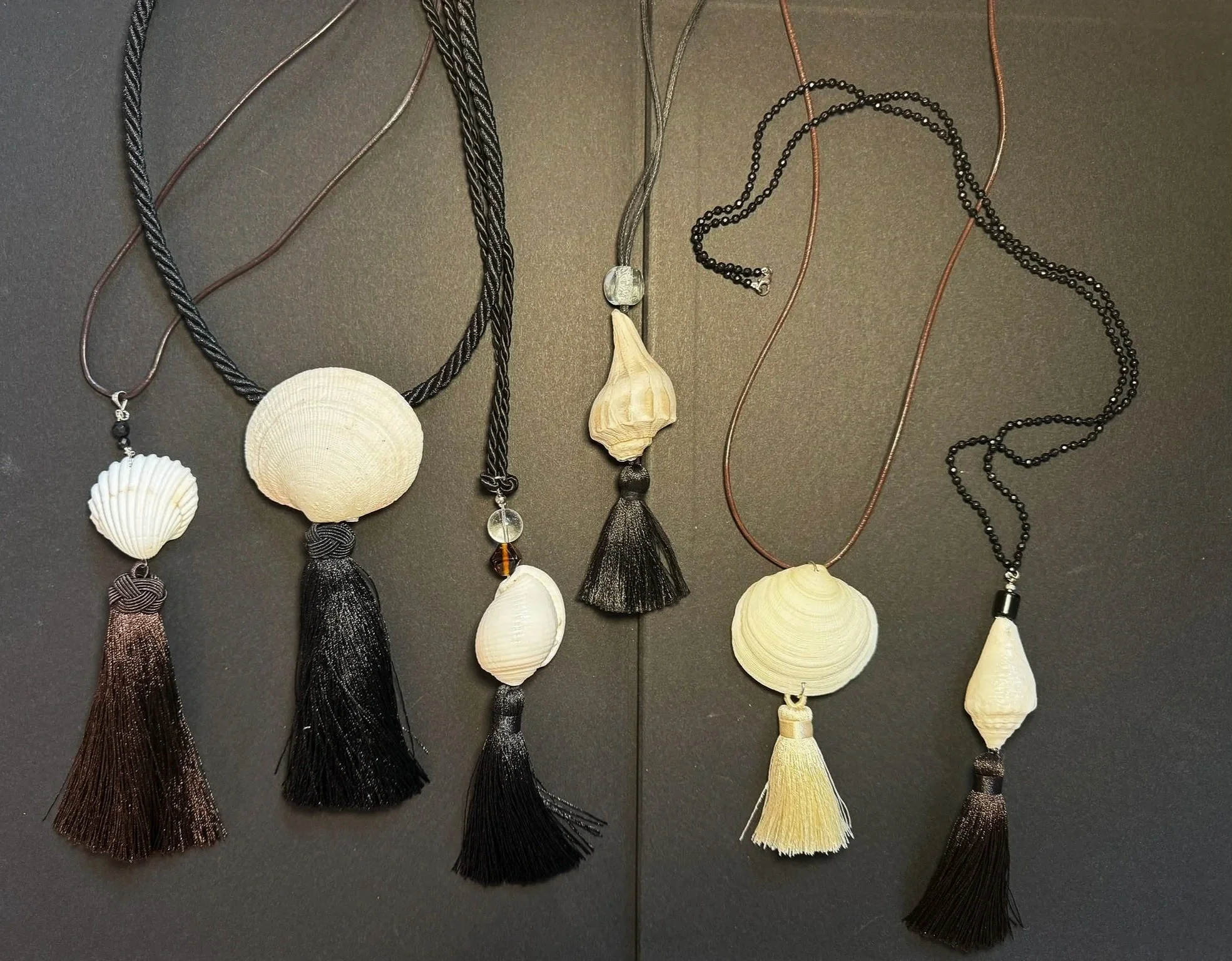 Collection of six necklaces with shell pendants and tassels on a black background.
