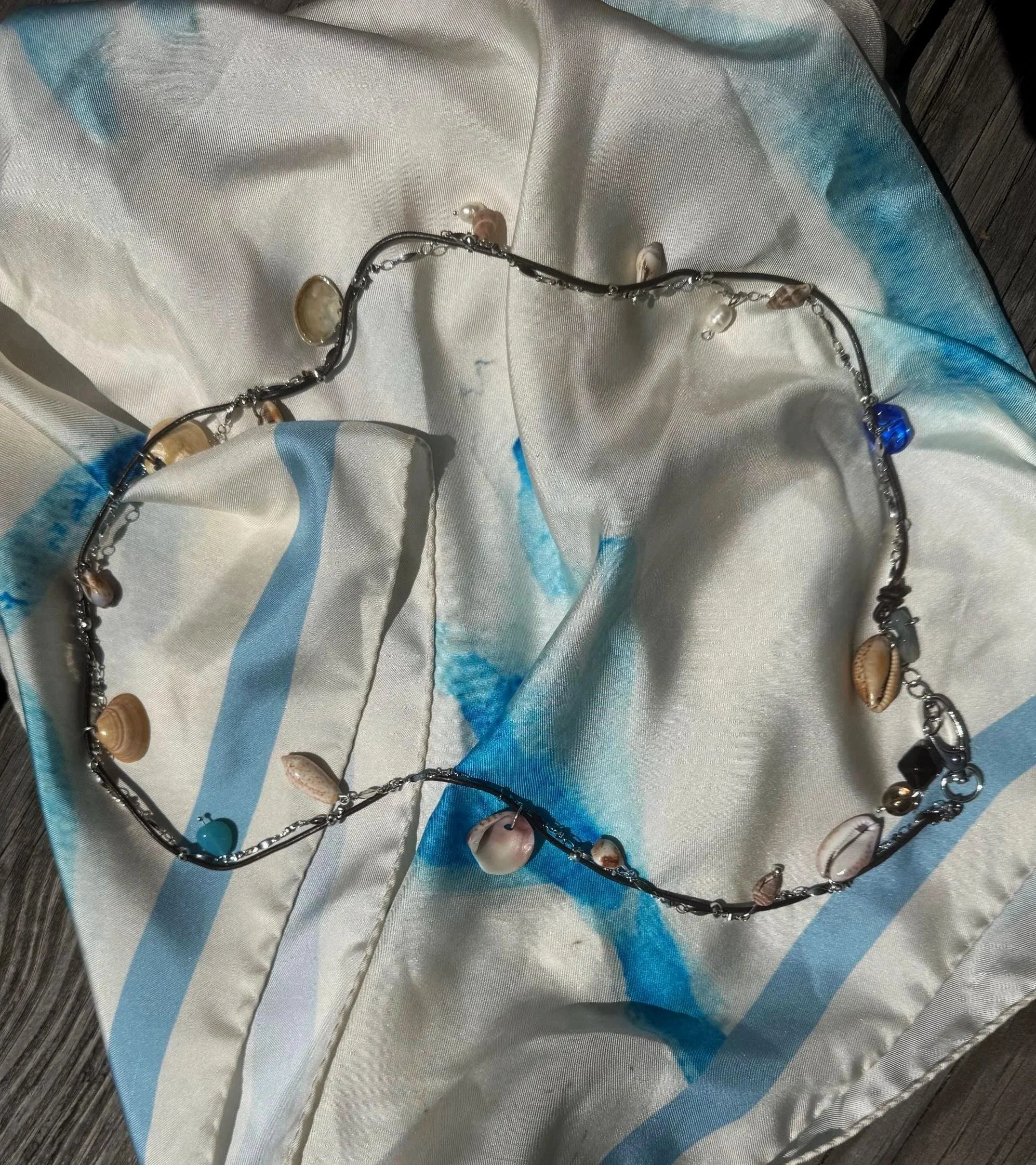 A seashell necklace with various shells and beads, laid out on a satin fabric with blue and white patterns.
