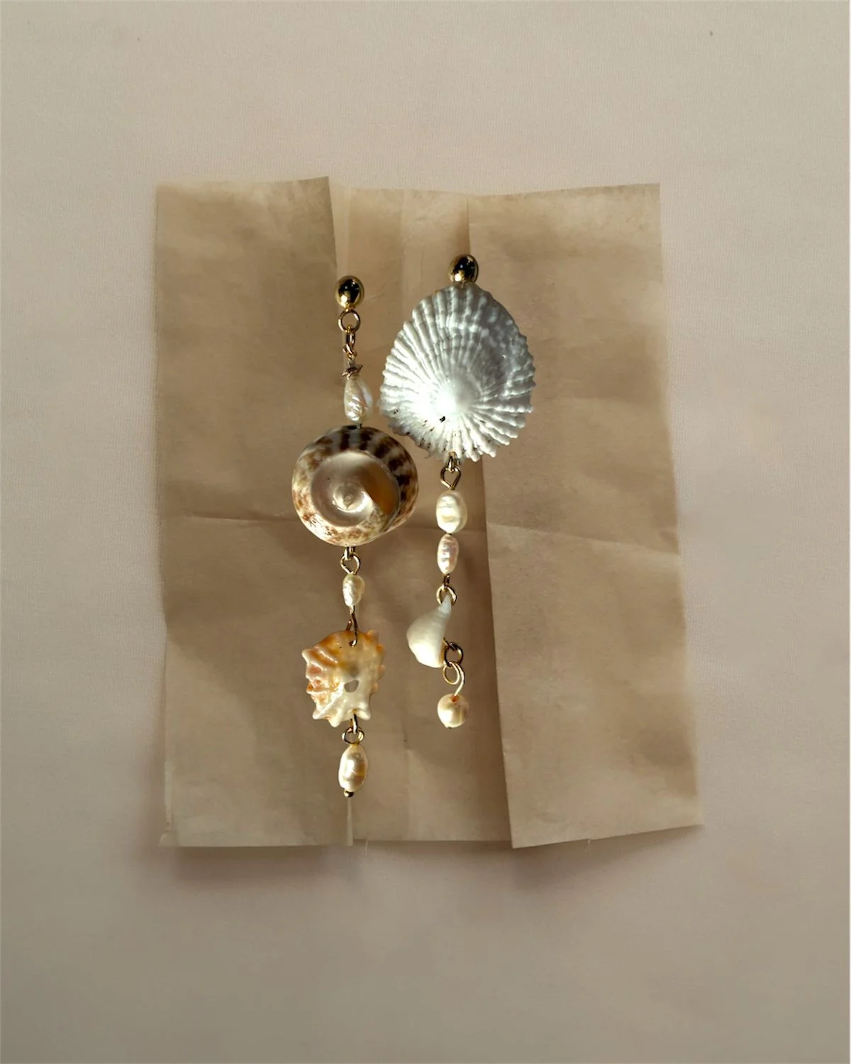 A pair of earrings featuring seashell designs and pearl accents, displayed against cream-colored paper.