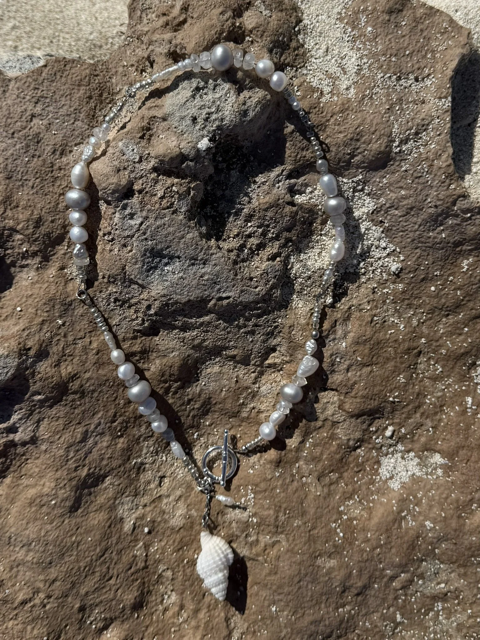 A pearl necklace with a shell pendant lying on a rough, brown rock surface.