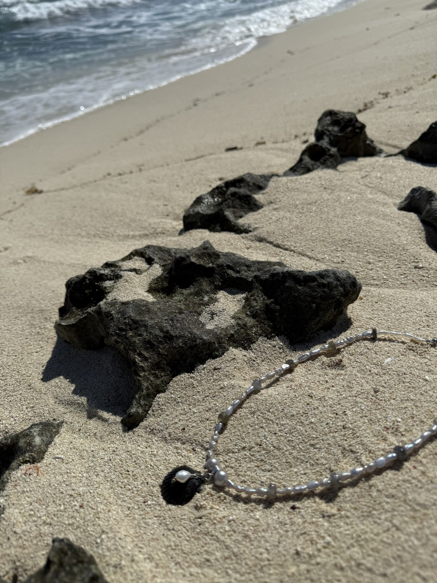 A jewelry necklace with pearls and a black shell pendant lies on sandy beach near black volcanic rocks, with the ocean and waves in the background.