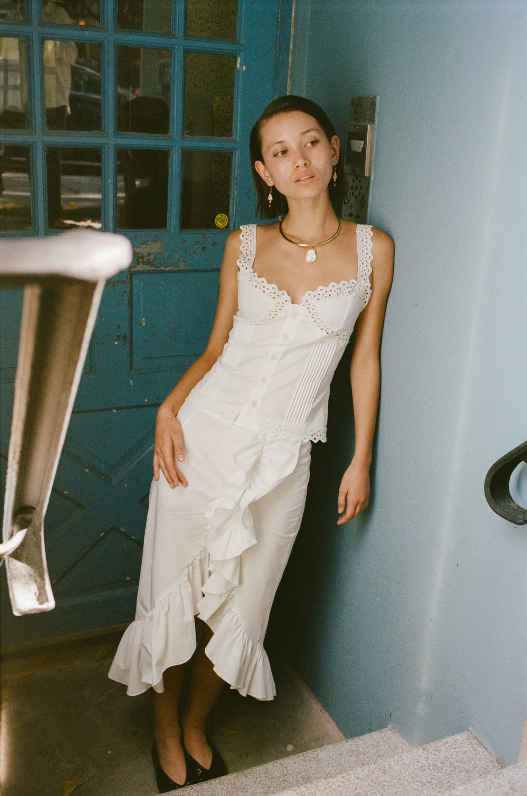 A young woman with short dark hair and light skin tone wearing a white dress, standing in an entryway near a blue door, with her back against the wall and looking slightly to the side.