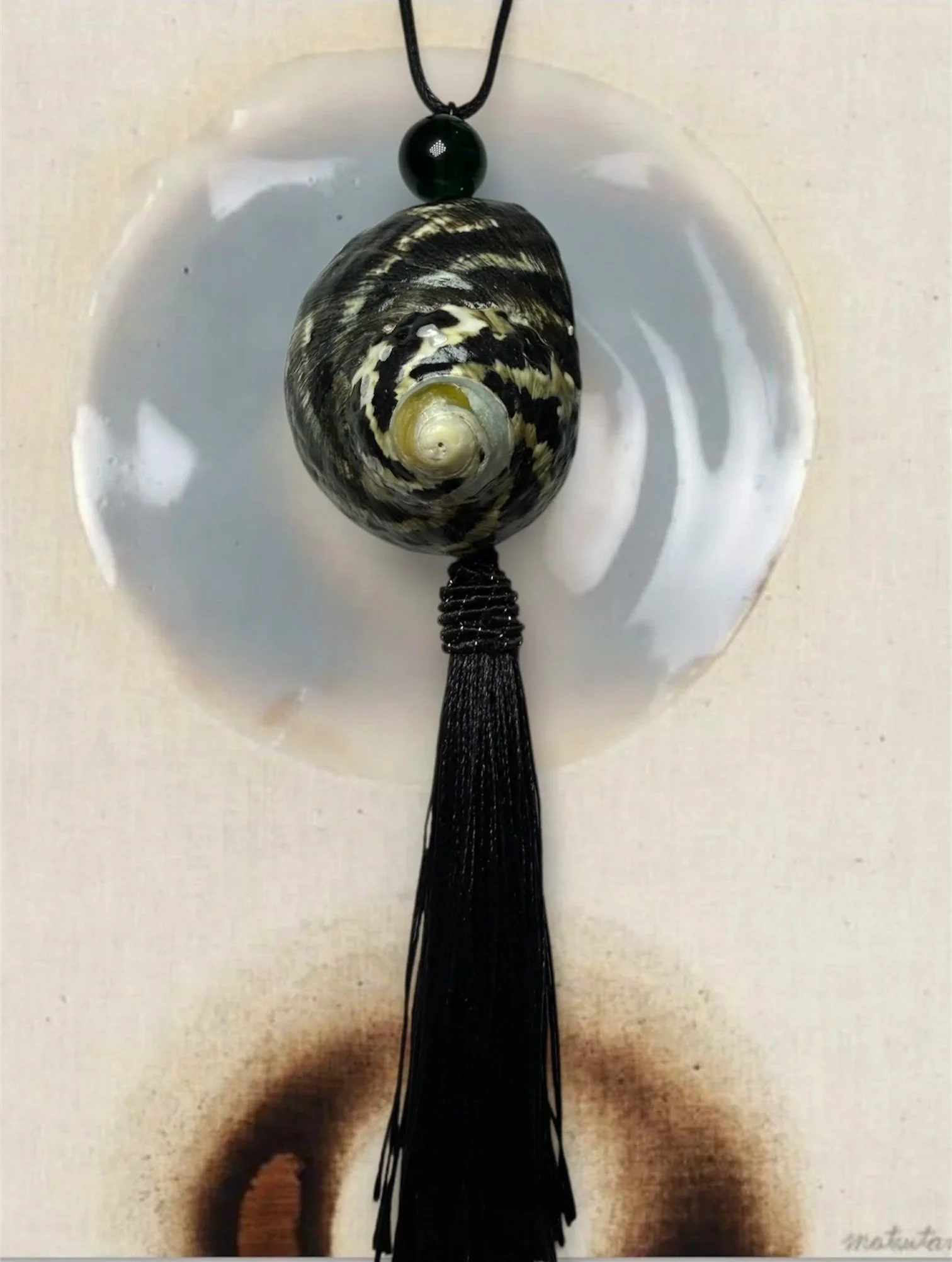 A pendant necklace featuring a shell with black and white patterns, a black round bead above the shell, and a long black tassel hanging below, displayed on a white surface.