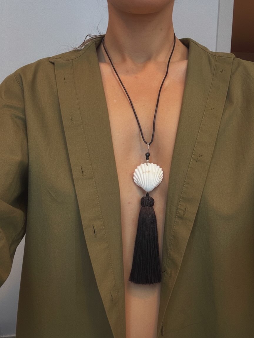 Person wearing an olive green blazer with an ocean shell pendant necklace and a black tassel hanging from the shell.