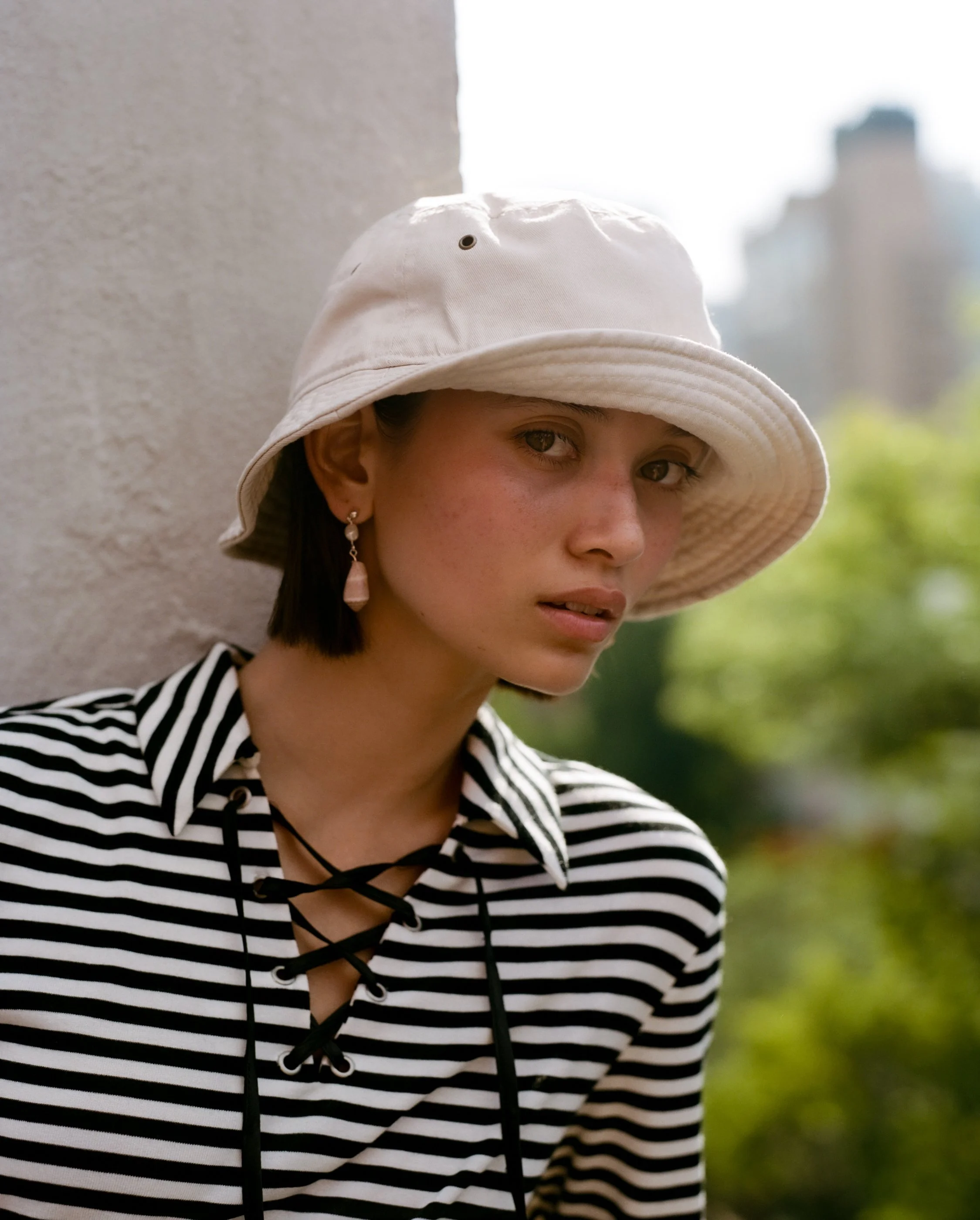 A young woman with short dark hair wearing a cream bucket hat, a black and white striped top with lace-up neckline, and dangling earrings, standing outdoors near a concrete wall with a blurred background of trees and buildings.