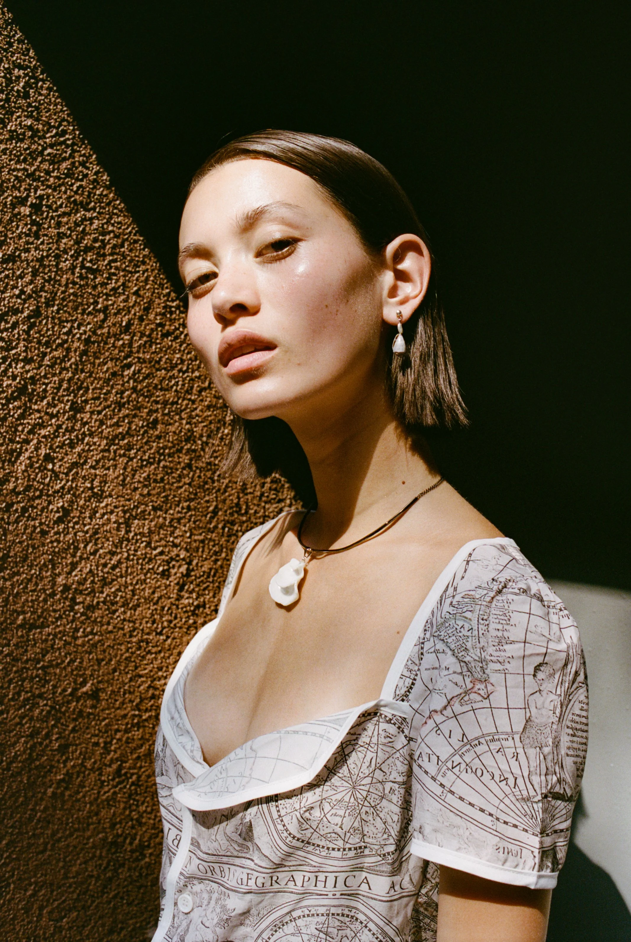 A young woman with short dark hair, wearing a necklace with a large pearl pendant and matching earrings, poses against a textured brown wall and a black backdrop. She is dressed in a white shirt with a nautical map print.