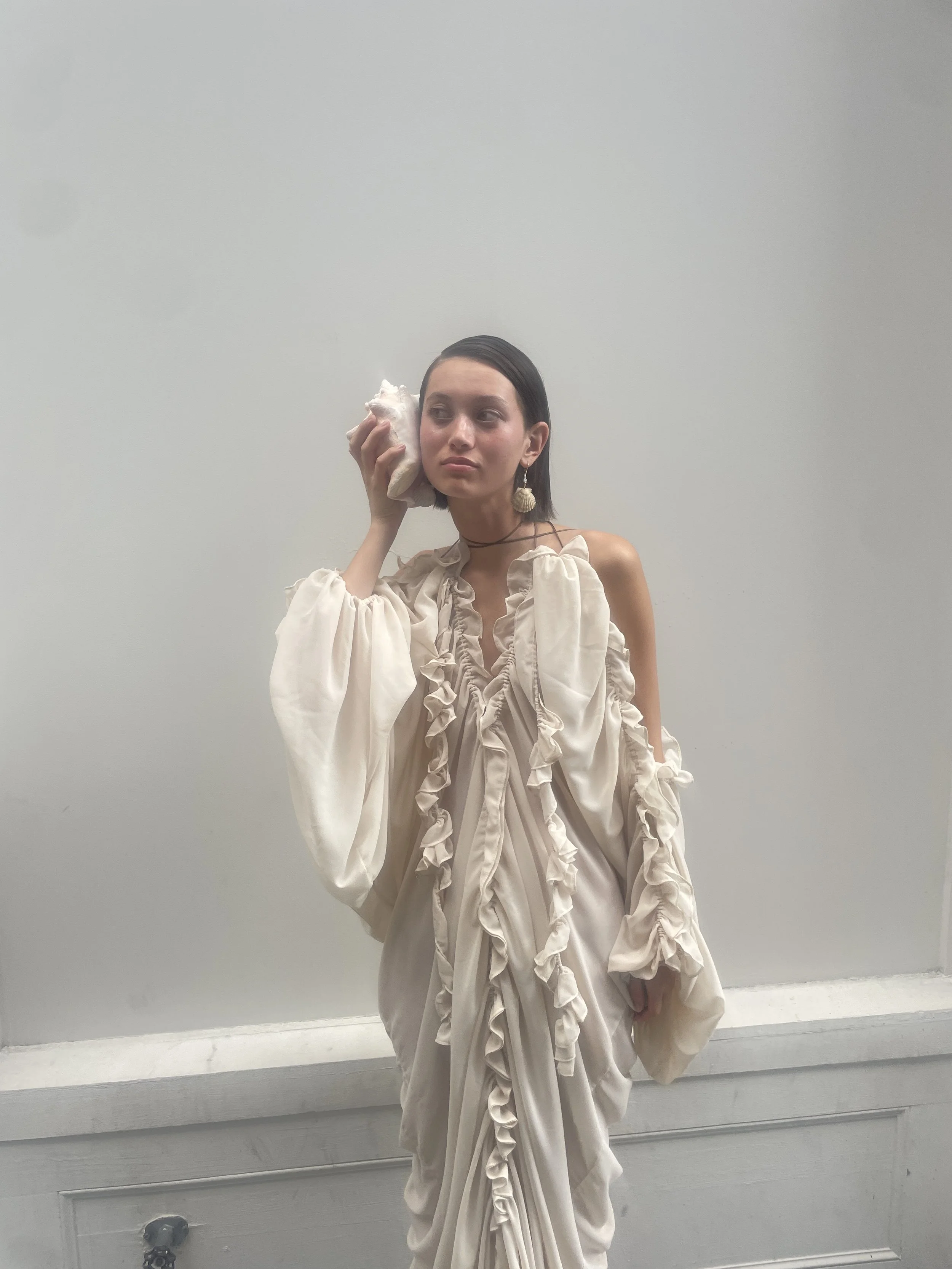 Young woman with dark hair, wearing an elegant ruffled beige gown, holding a shell near her face, standing against a plain white wall.