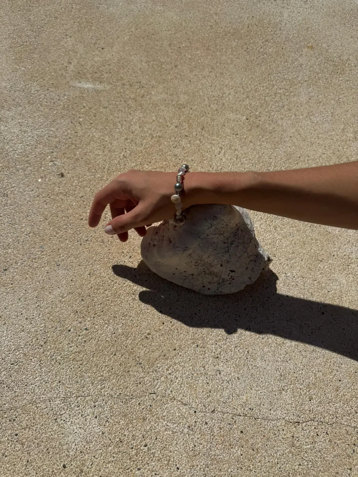 A hand wearing a pearl bracelet rests on a large, textured white shell on sandy ground, casting a shadow.