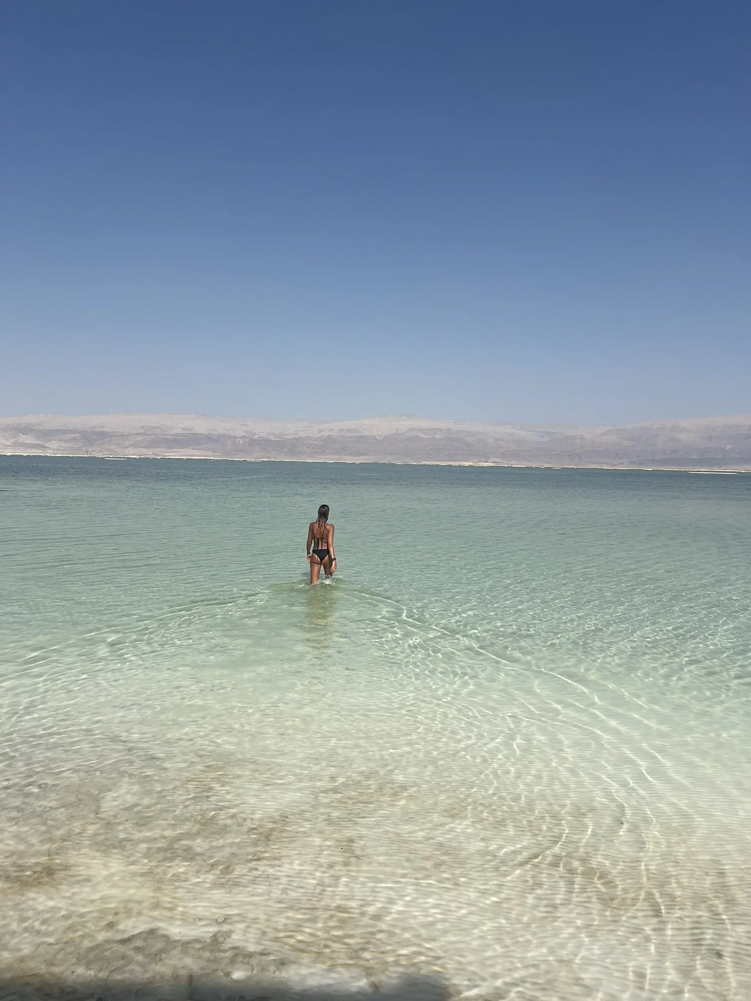 A woman in a black swimsuit wading into clear, shallow water at the beach under a blue sky.