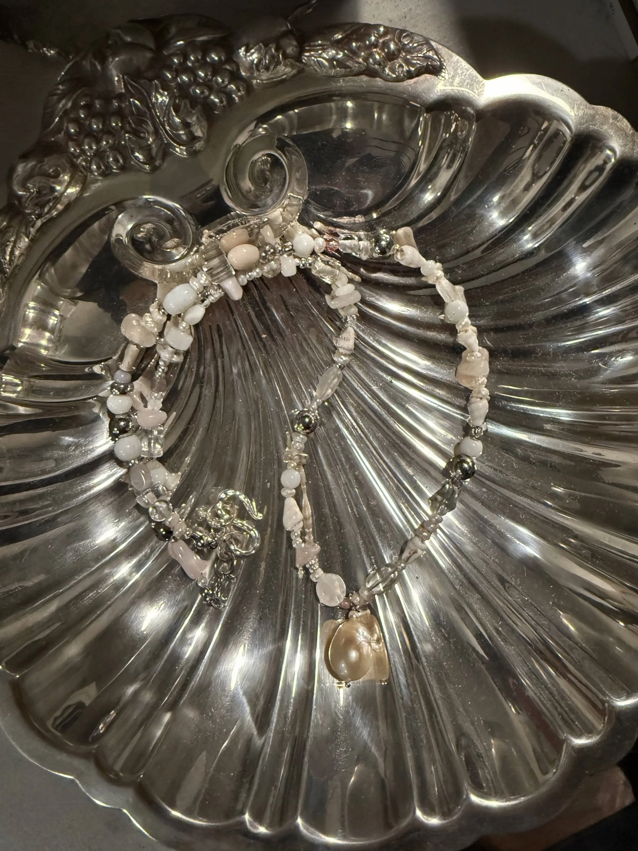 A pearl and beaded necklace with silver and clear beads, and a large pearl pendant, resting on an ornate silver dish with floral and fruit embellishments.