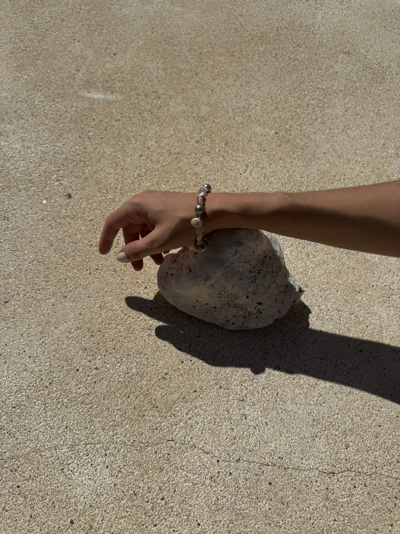 A hand wearing a beaded bracelet resting on a large sea shell on a sandy surface, casting a shadow.