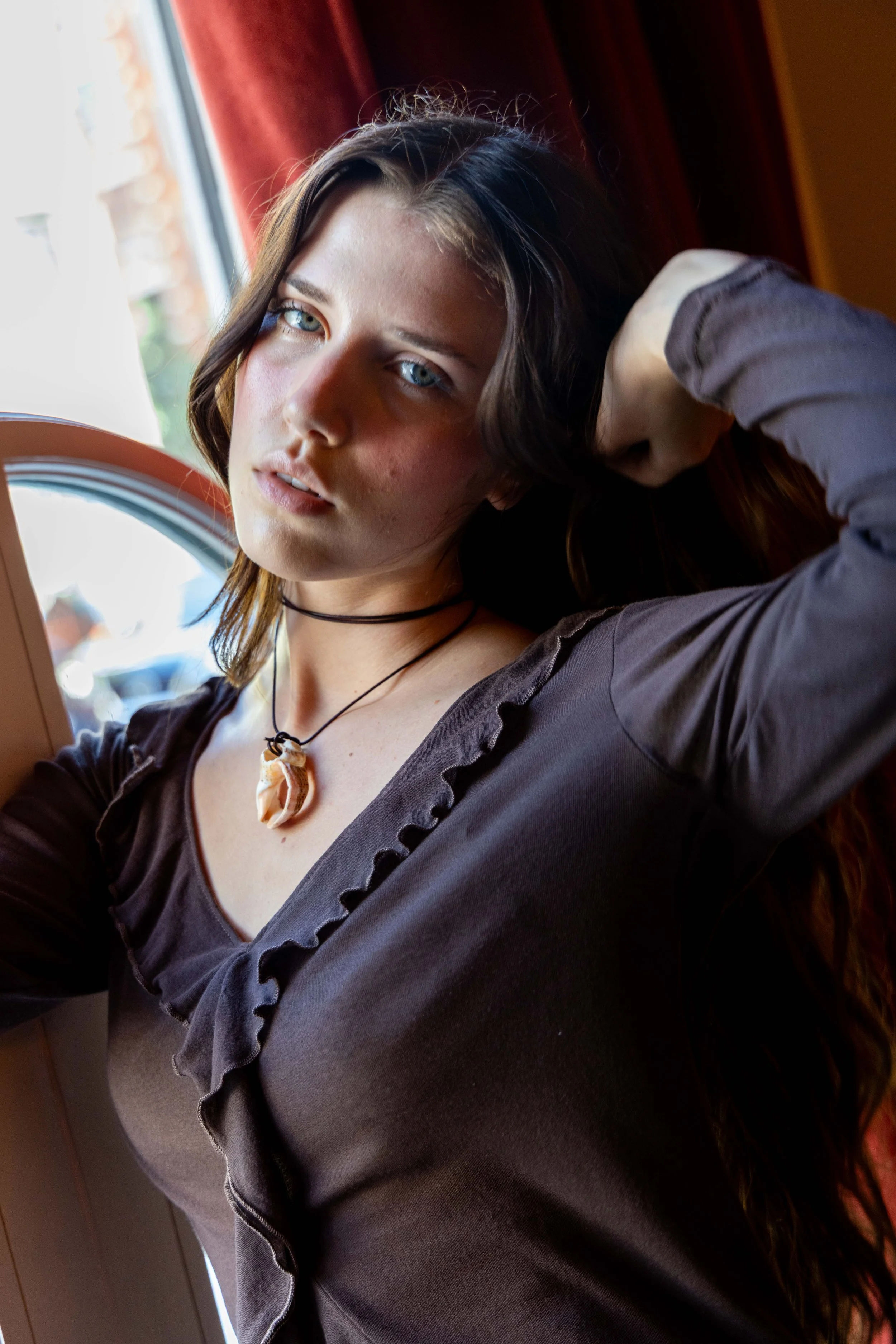 Young woman with brown hair and blue eyes sitting near a window, wearing a dark top and a shell necklace, gazing at the camera.