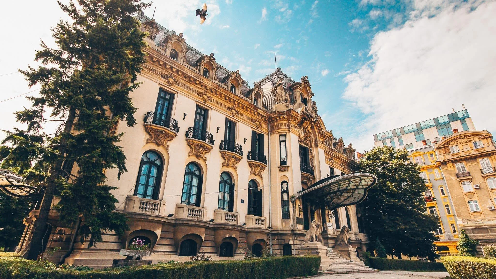 A historic building with ornate architecture, large windows, and decorative statues on the roof, situated next to a modern glass building, with trees and a clear sky in the background.