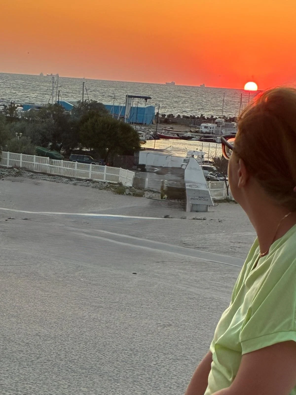 A woman with glasses and reddish hair looking towards the sunset over the ocean, with a road and marina in the foreground.
