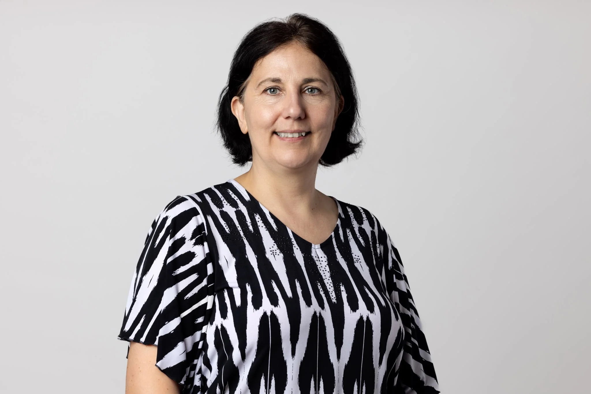 A woman with short black hair and blue eyes, smiling, wearing a black and white patterned top, against a neutral gray background.