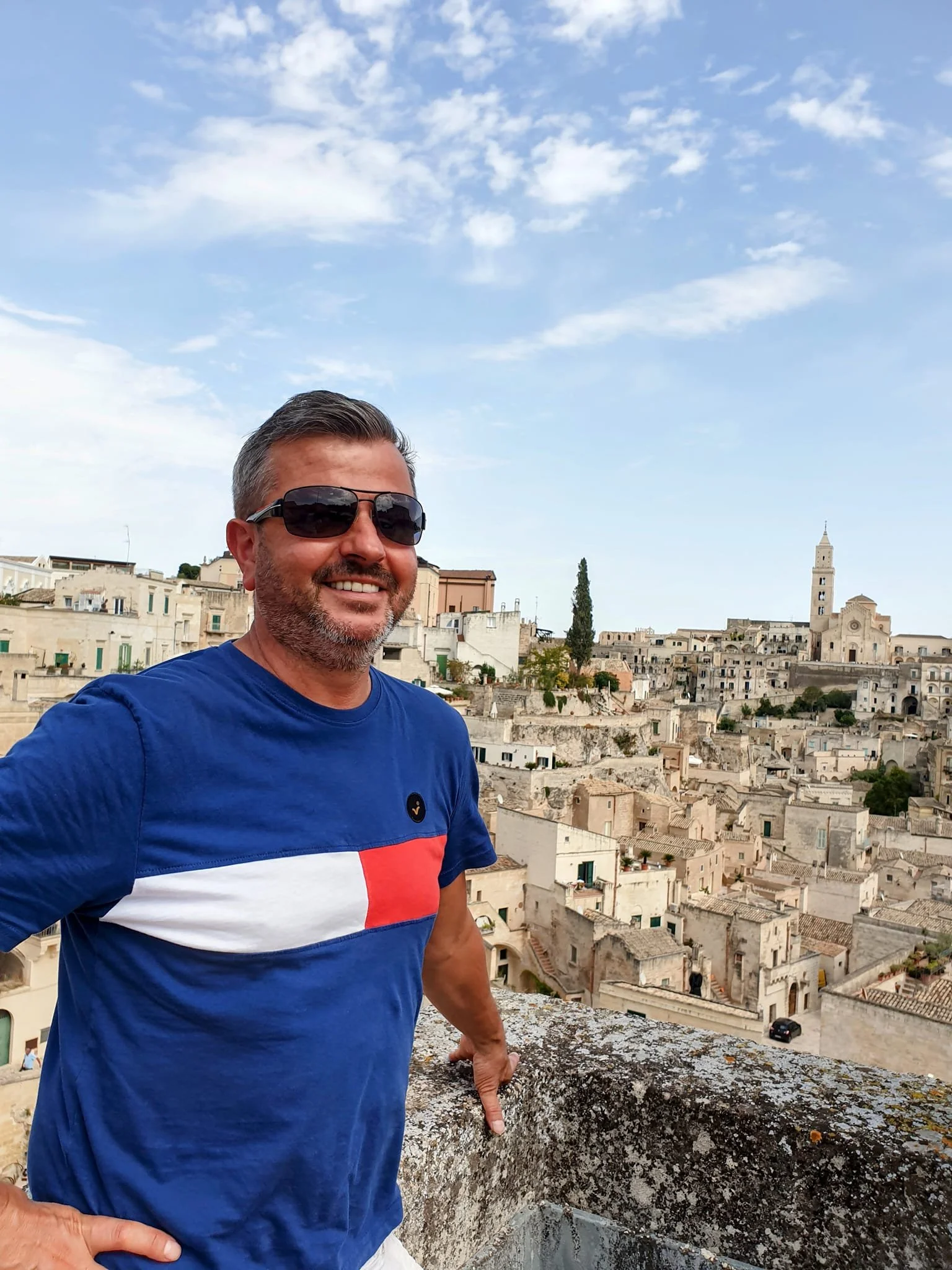 A man with dark hair, beard, and sunglasses smiling while standing in front of a cityscape with historic buildings and a tall church tower under a partly cloudy sky.