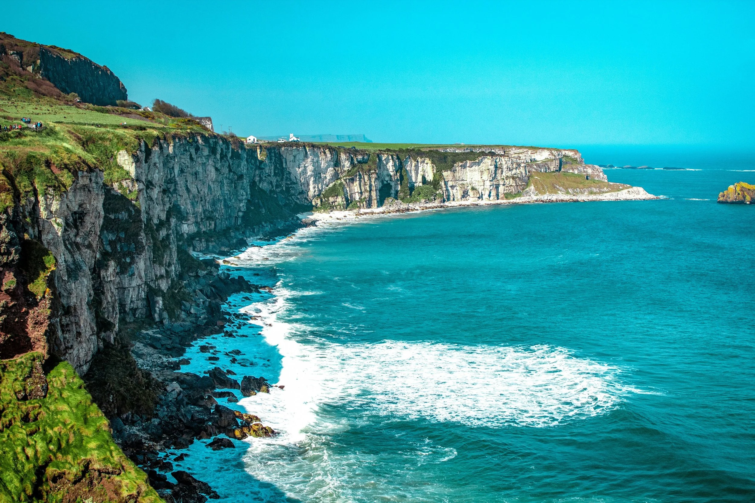 Cliffs along the coast with green grassy tops, rocky faces, and a bright blue ocean below. A few small buildings and pathways are visible on the grassy areas.