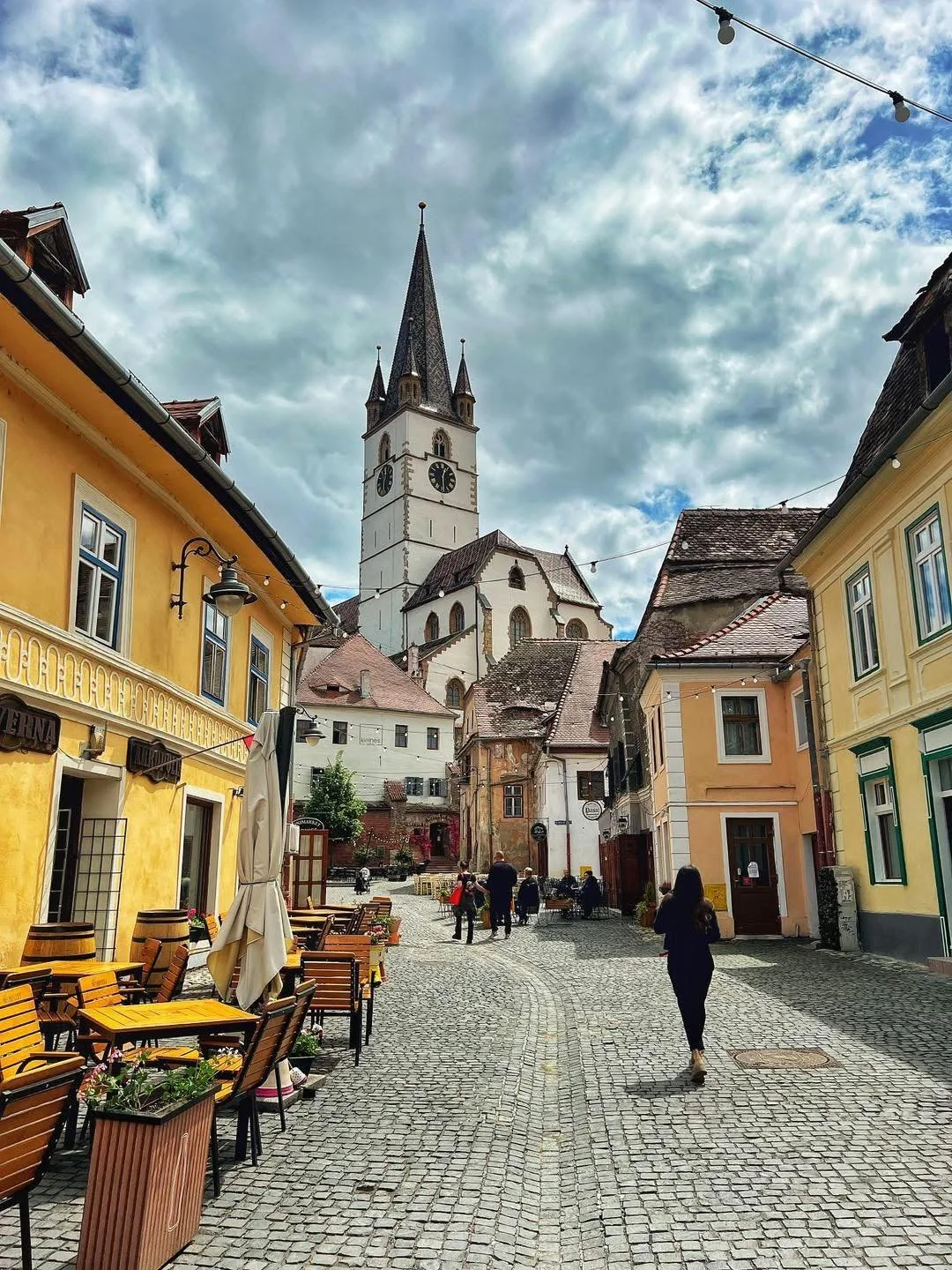 A cobblestone street in a European town with colorful buildings and outdoor seating, with a large church steeple in the background under a partly cloudy sky.