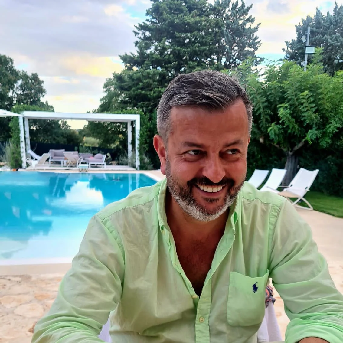 A man smiling and sitting outdoors near a swimming pool with lounge chairs and greenery in the background.