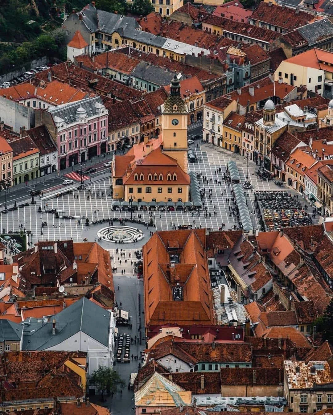 An aerial view of a European city square with red-tiled roofs, pastel-colored buildings, a central clock tower, and a circular fountain, with people walking and parked cars around.