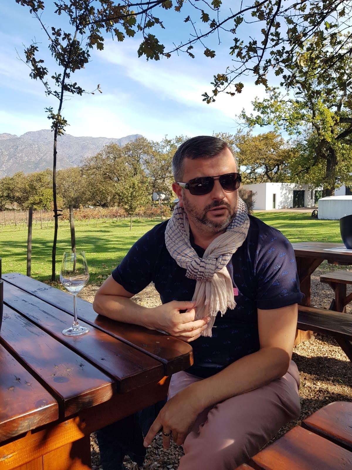 A man wearing sunglasses and a beige scarf sitting at a wooden picnic table outdoors in a rural setting with trees, grass, and mountains in the background.