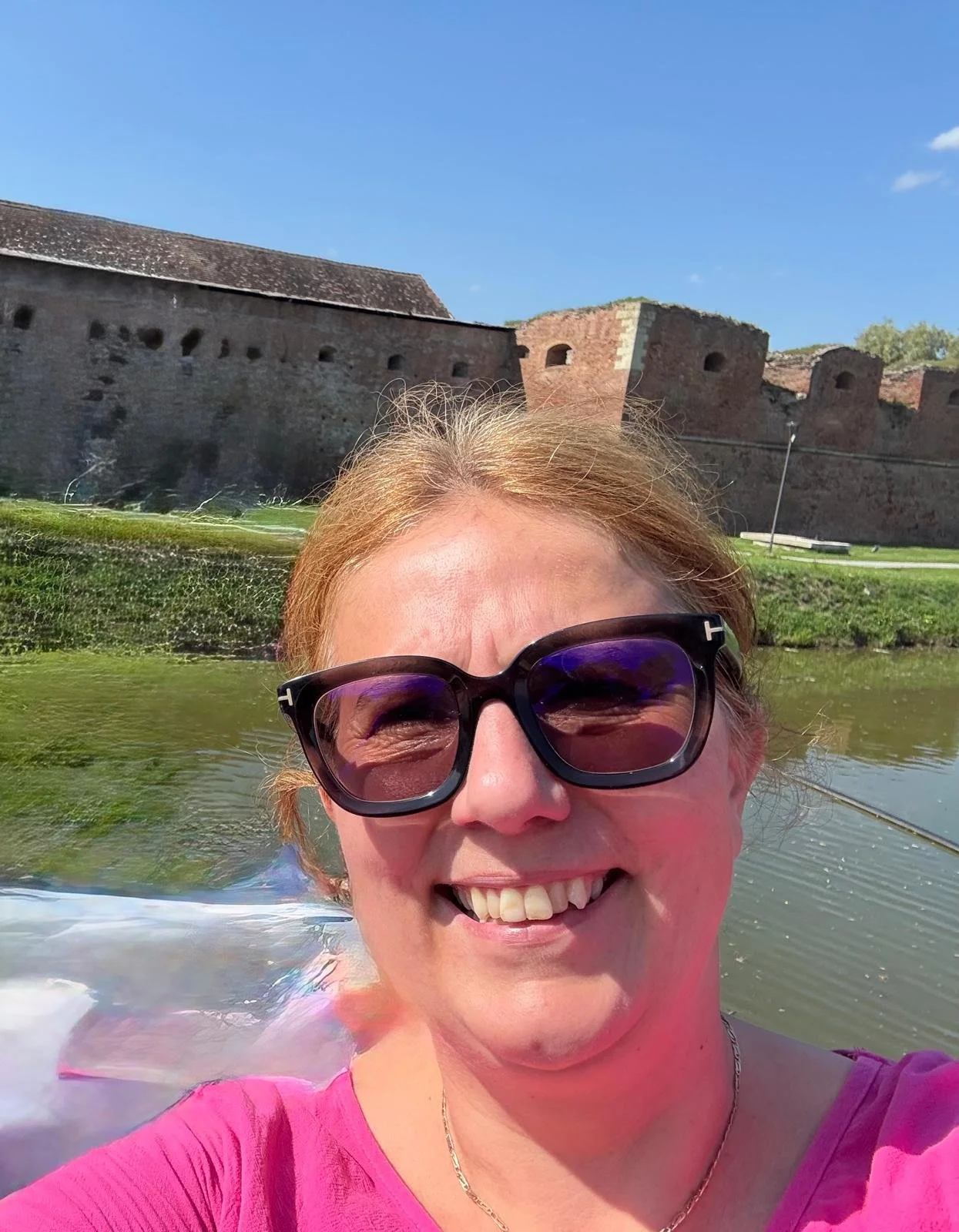 A smiling woman wearing sunglasses and a pink top taking a selfie in a boat on a river, with a historic stone fortress in the background under a clear blue sky.