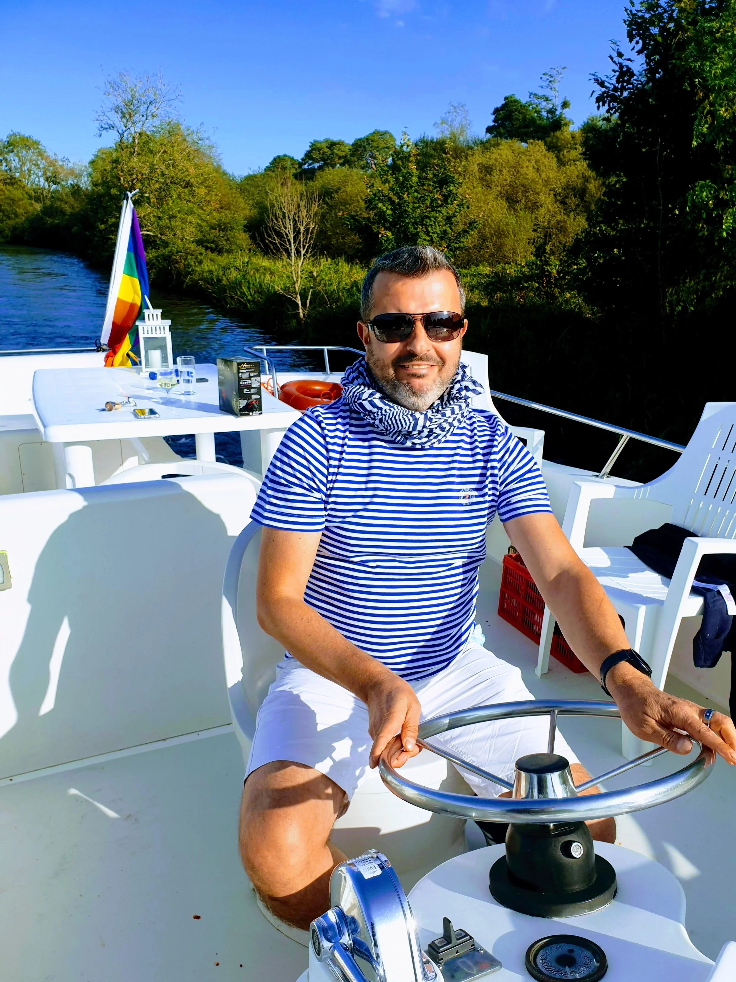 A man in a blue and white striped shirt and white shorts is smiling while sailing on a boat in a river or lake surrounded by trees, wearing sunglasses and a neck scarf.