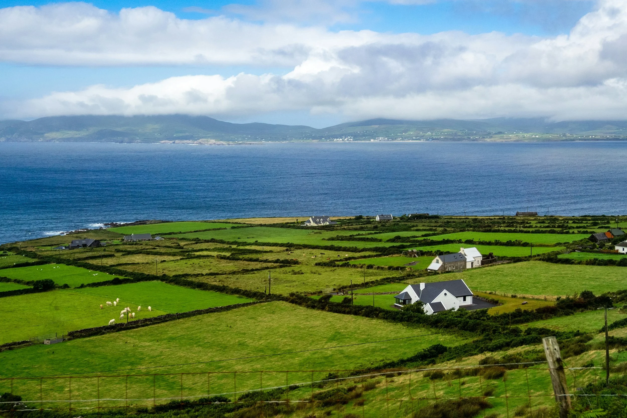 Green fields divided by stone walls with a few white houses, near water under a partly cloudy sky.