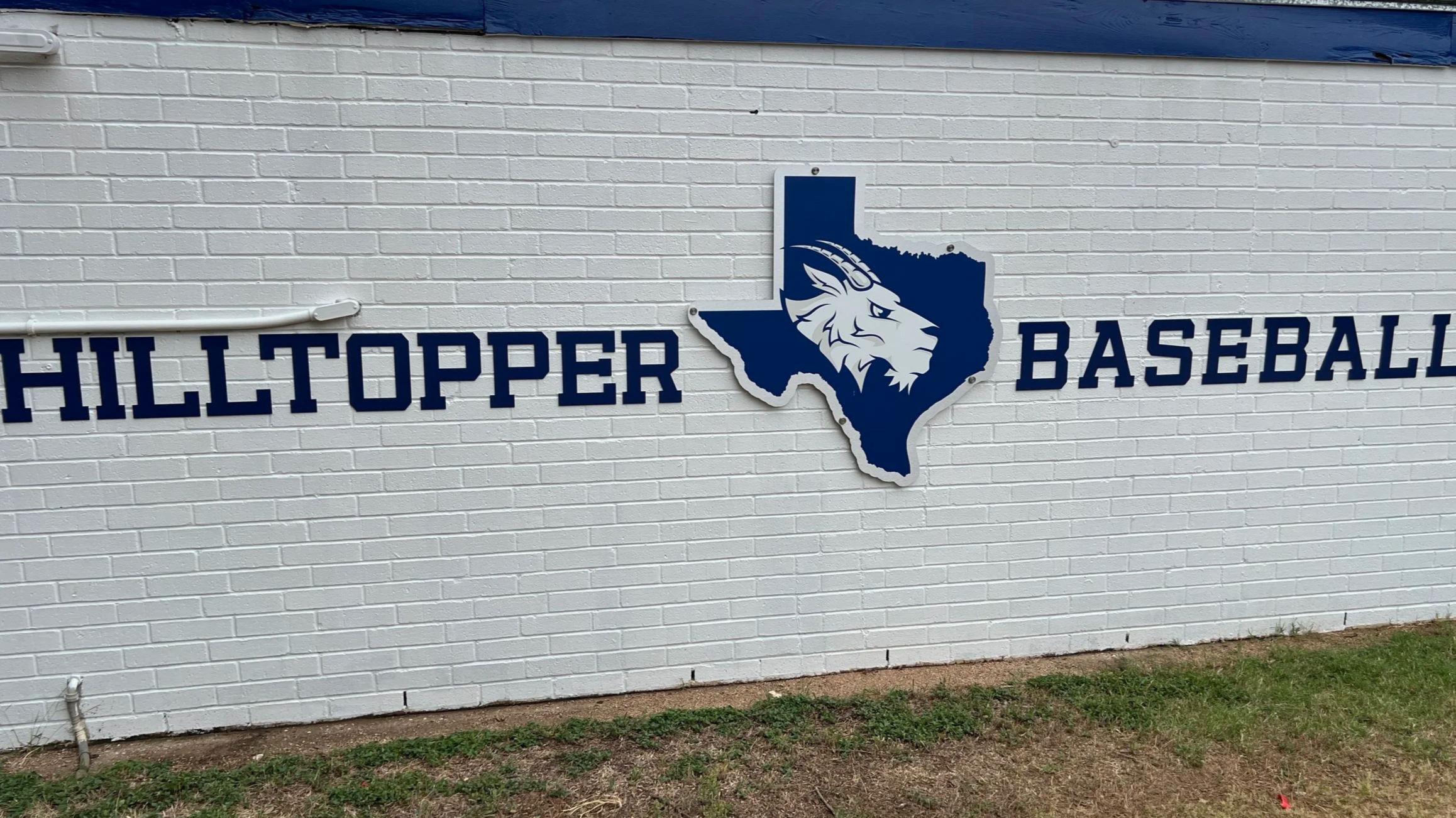 A sign on a white brick wall reads 'HILLTOPPER BASEBALL' with a blue outline of Texas that contains a lion's head logo.