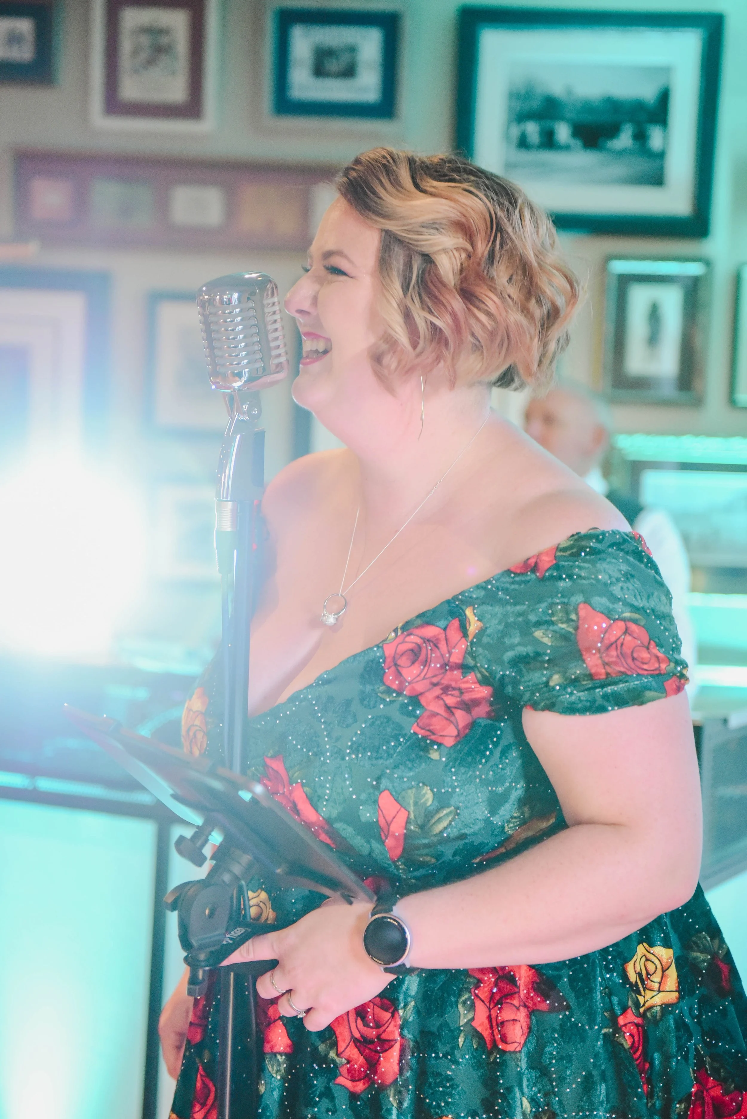 A woman singing into a vintage microphone at a music event, with framed photos on the wall in the background.