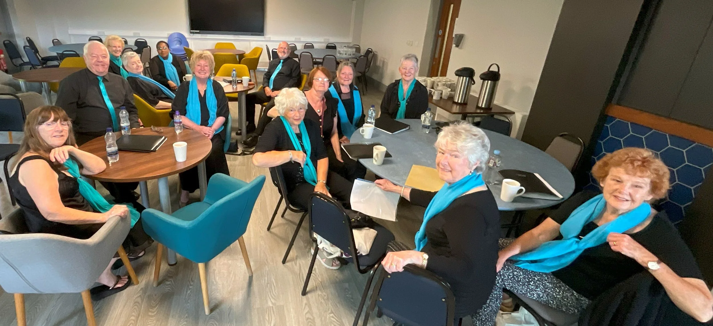 Group of elderly people sitting and standing around tables in a conference or meeting room, smiling, with beverages and documents on the tables, wearing black clothing with turquoise scarves or ties.