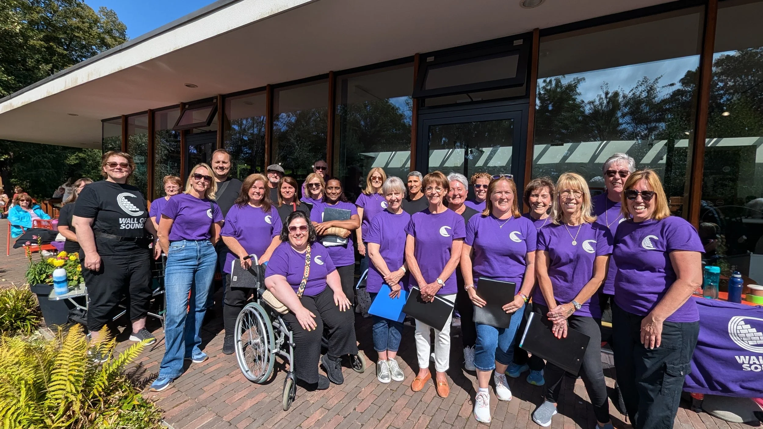 Group of people standing outdoors in front of a building, some wearing purple shirts with a logo, smiling for a group photo.